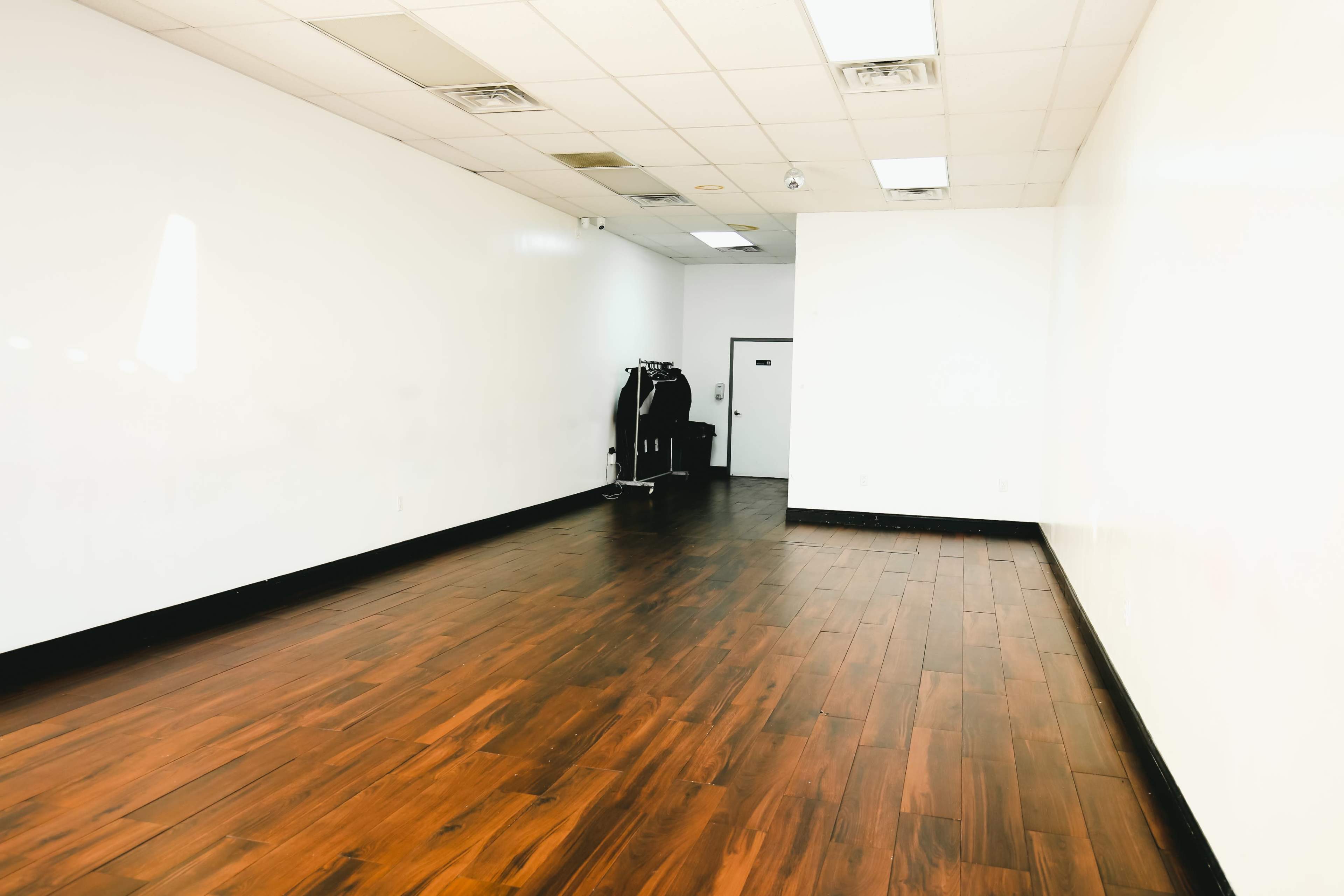 The image shows a spacious, empty room with wooden flooring and white walls, featuring a dark rack in the corner.