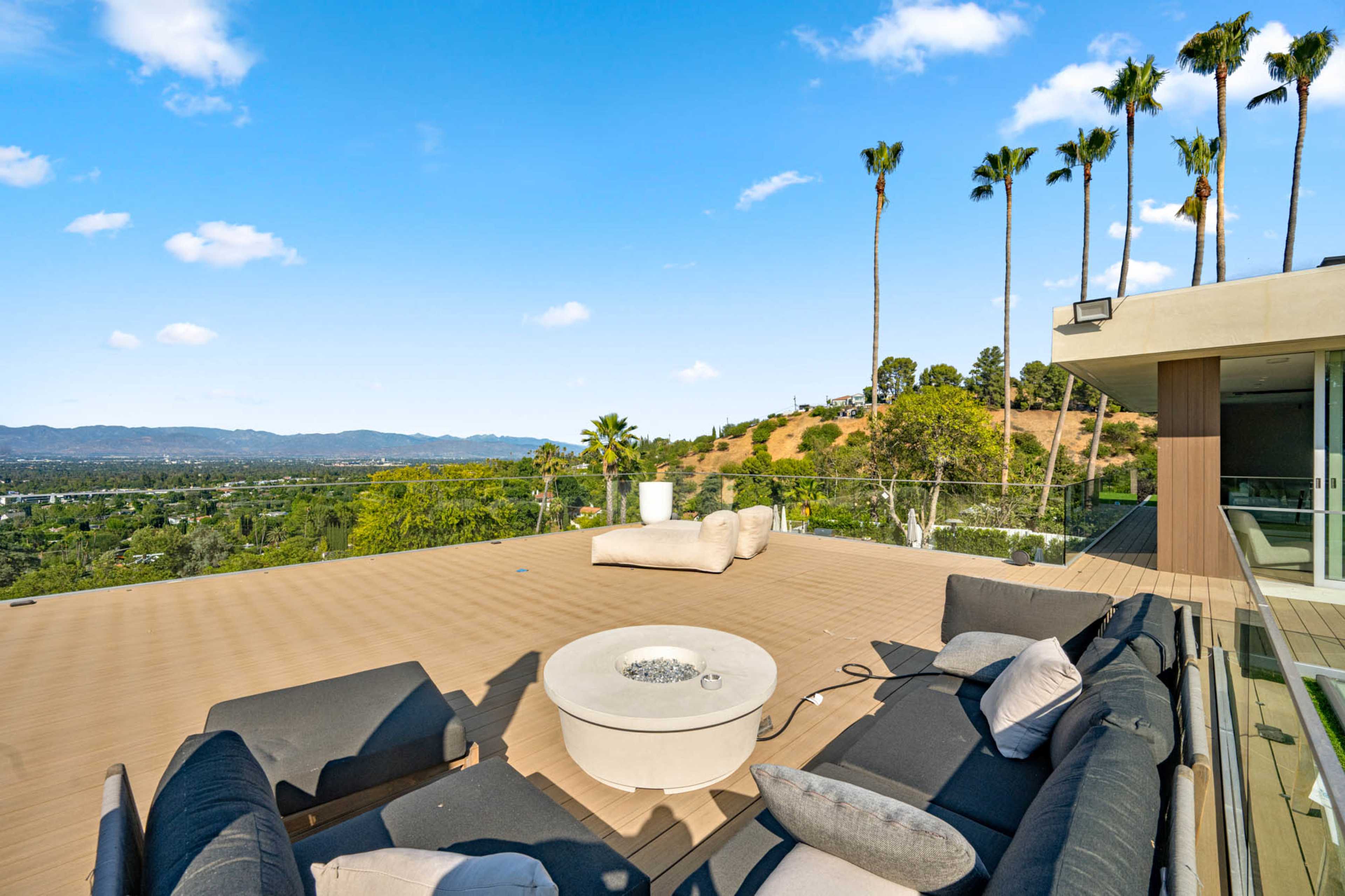 The image shows a spacious outdoor deck with modern seating and a fire pit, surrounded by palm trees and a scenic view of rolling hills and mountains.