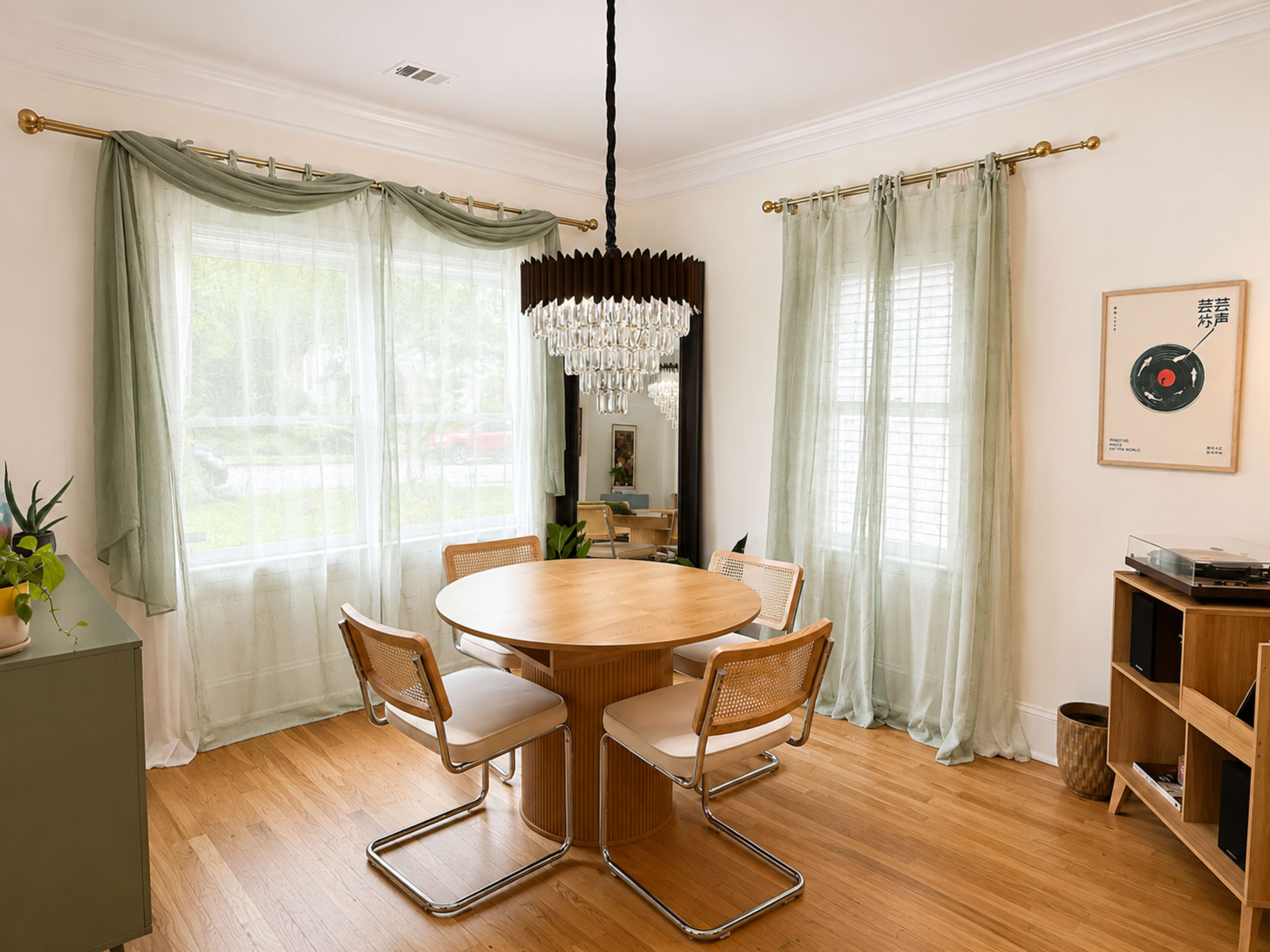 The image depicts a dining area with a round wooden table, four chairs, a chandelier, and large windows adorned with sheer curtains.