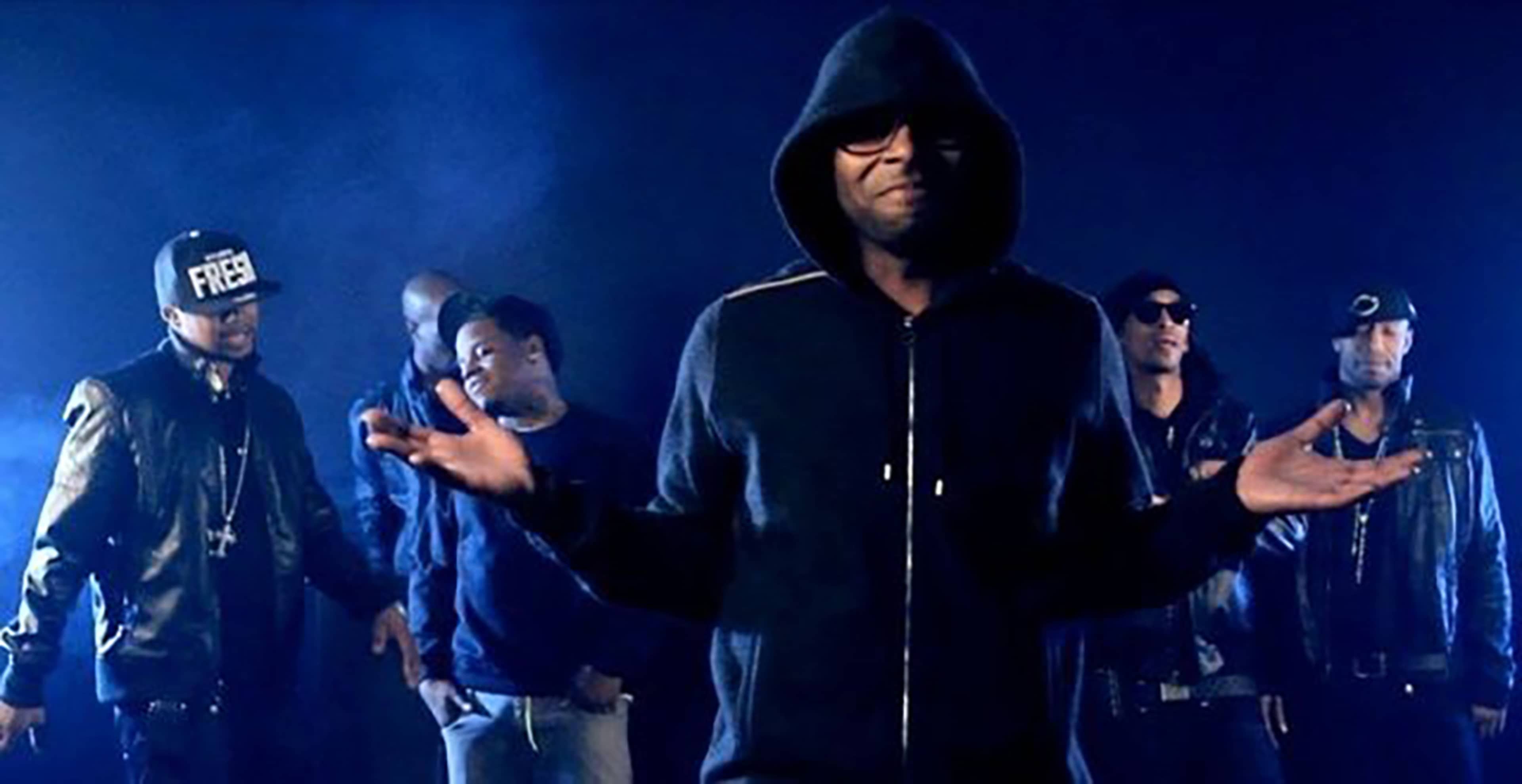 A group of men, dressed in dark clothing, stands in a spotlight against a blue backdrop, with one man in the foreground gesturing with his hands.