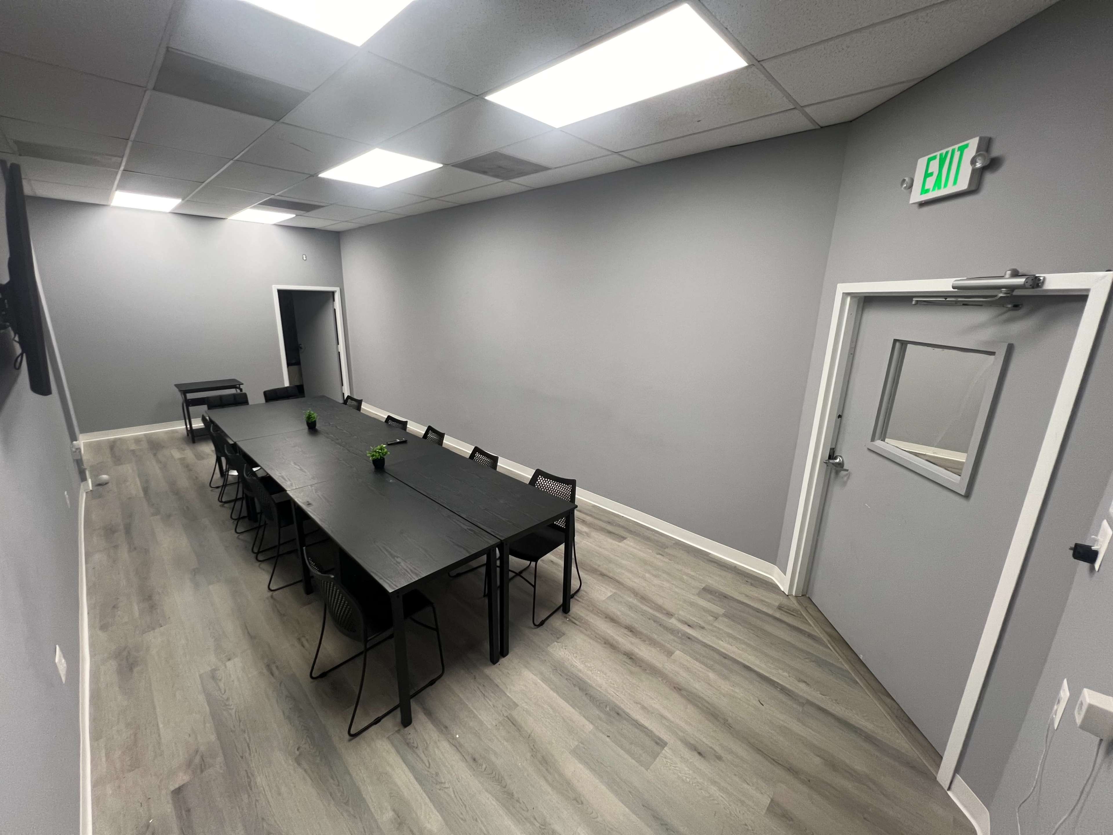 A sparse conference room featuring a long black table surrounded by chairs, with gray walls and a door on one side.