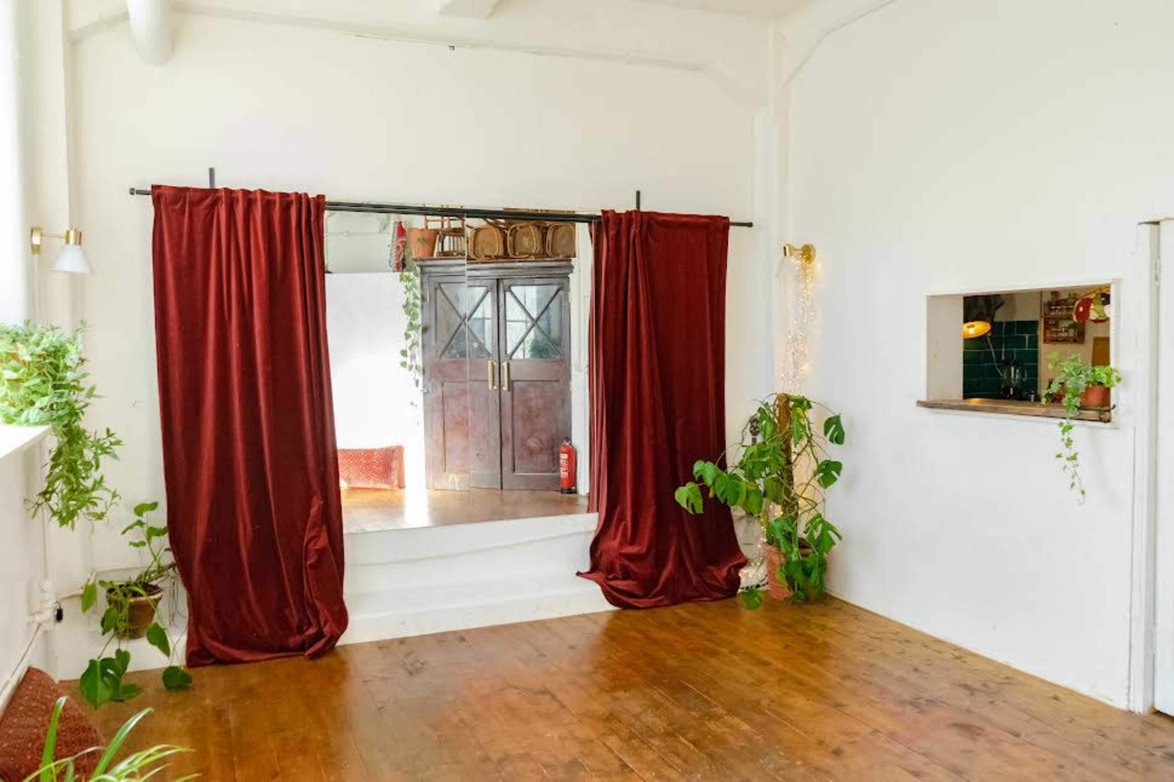 The image shows an interior space with wooden flooring, red curtains draped on a stage area, and a small kitchenette with plants in the corner.
