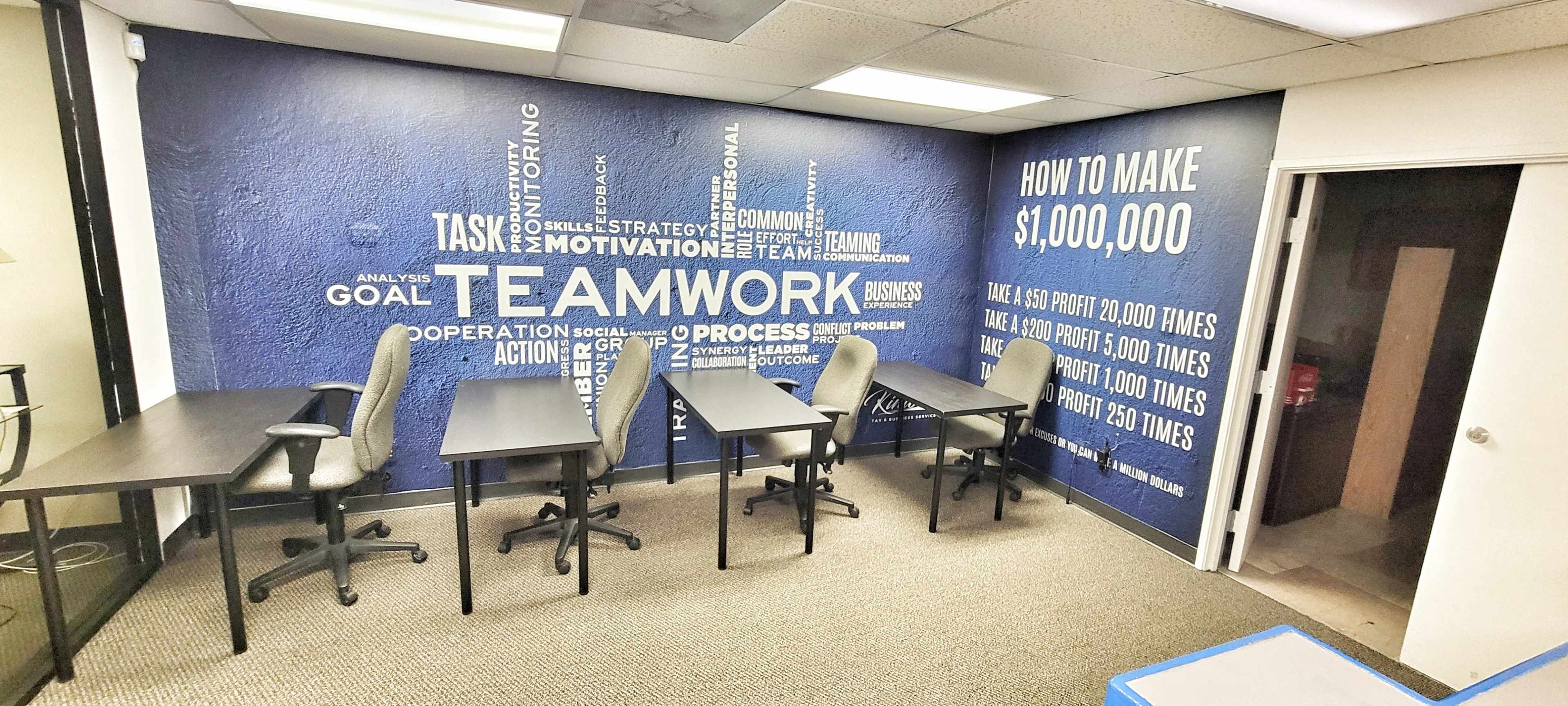 The image shows an office space featuring a blue wall with motivational phrases related to teamwork and business, along with several empty desks and chairs arranged in the room.