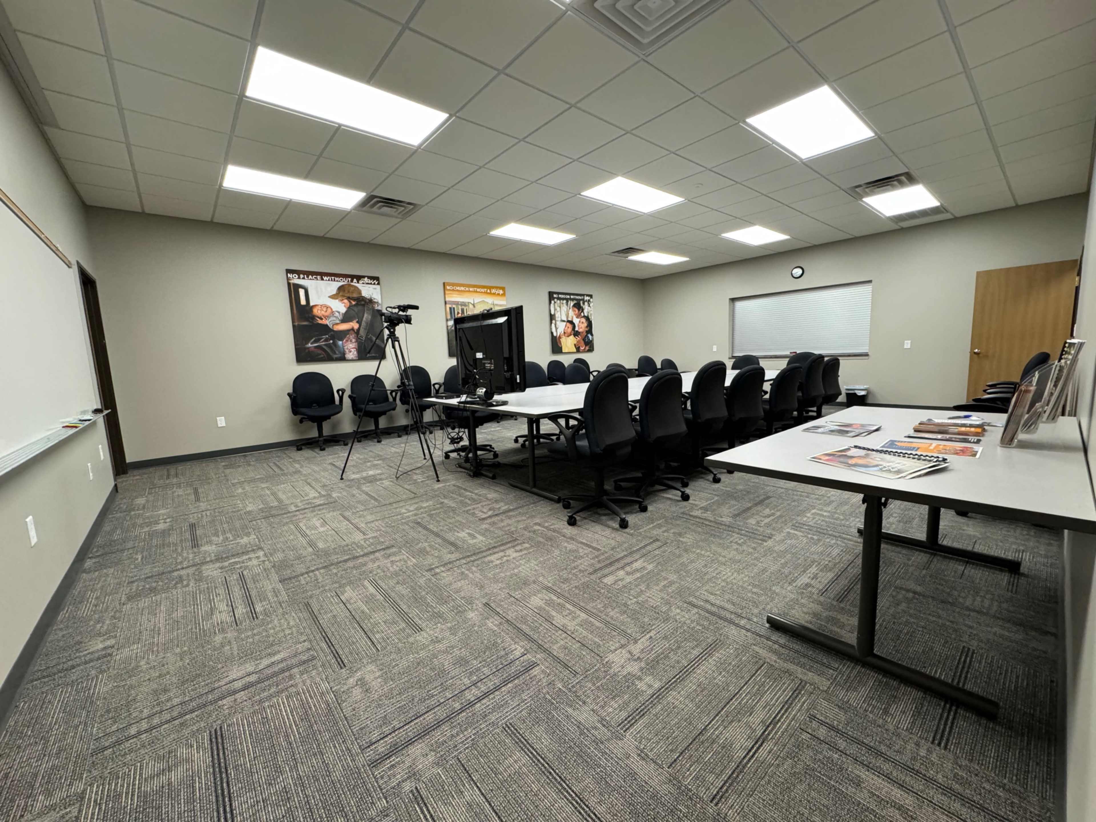 A conference room features a large table surrounded by chairs, with a whiteboard and a wall-mounted clock, and posters on the walls.