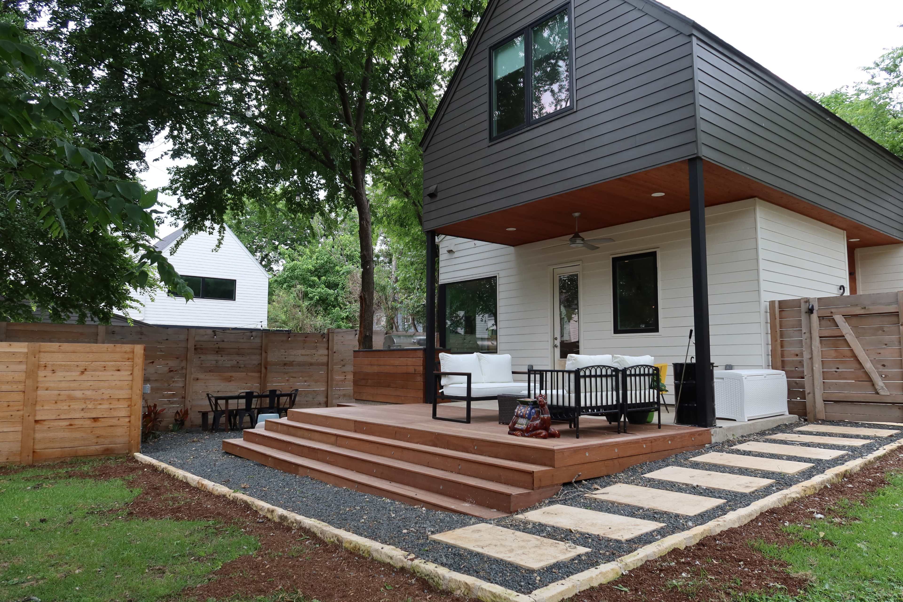 A modern house with a wooden deck and outdoor seating area, surrounded by a fenced yard and landscaping.