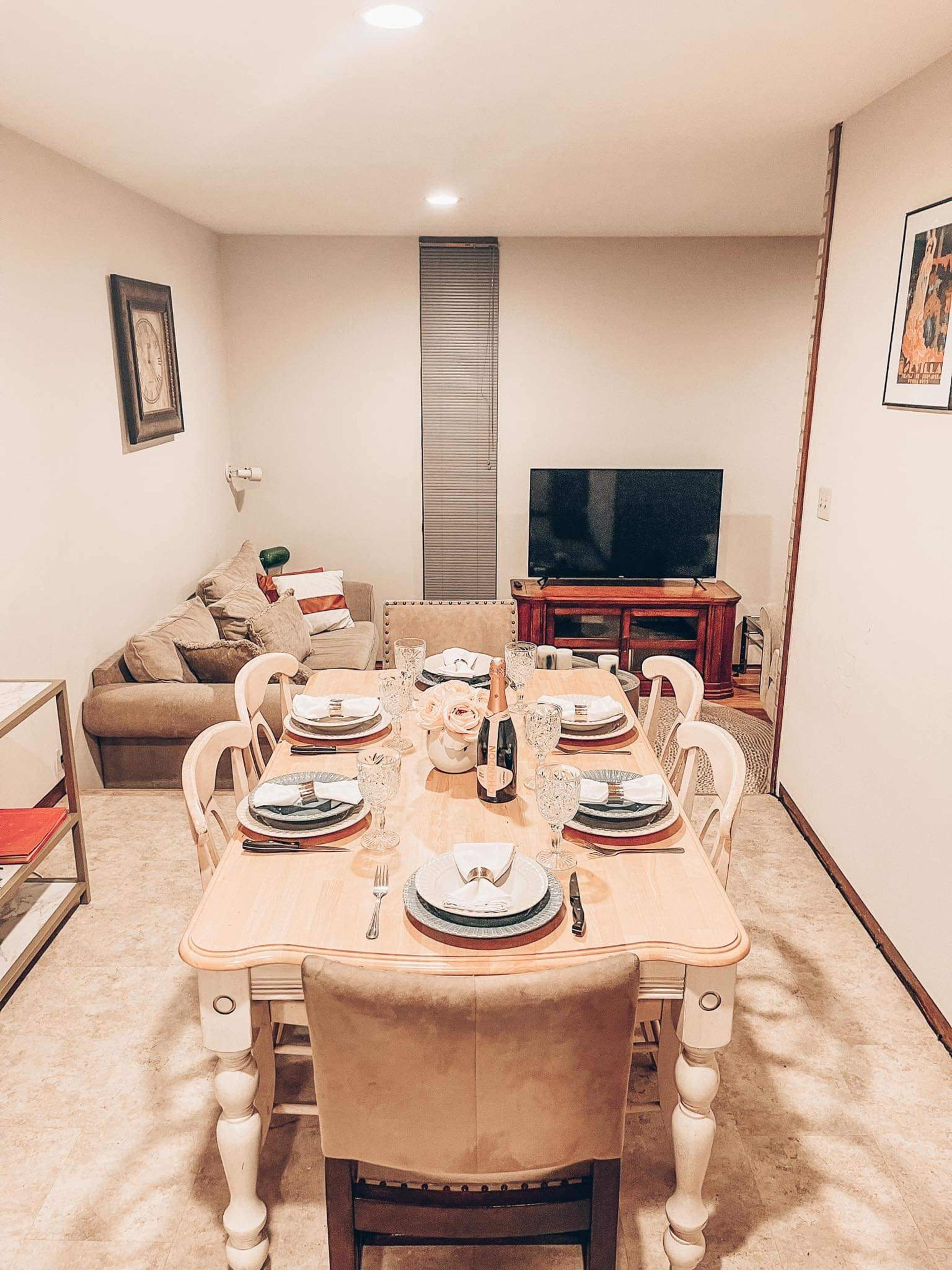 A dining area features a table set for eight with plates and glasses, a television, and a sofa in the background.