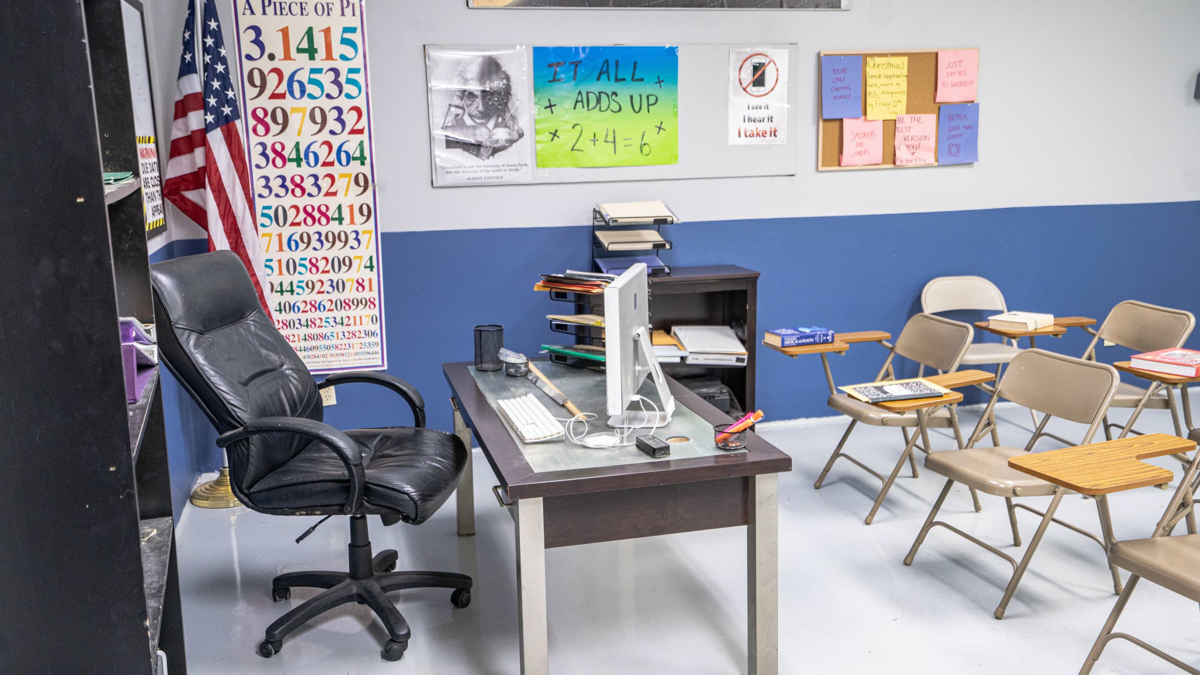 A classroom is set up with a desk and computer in front, chairs arranged for students, and educational materials displayed on the walls.