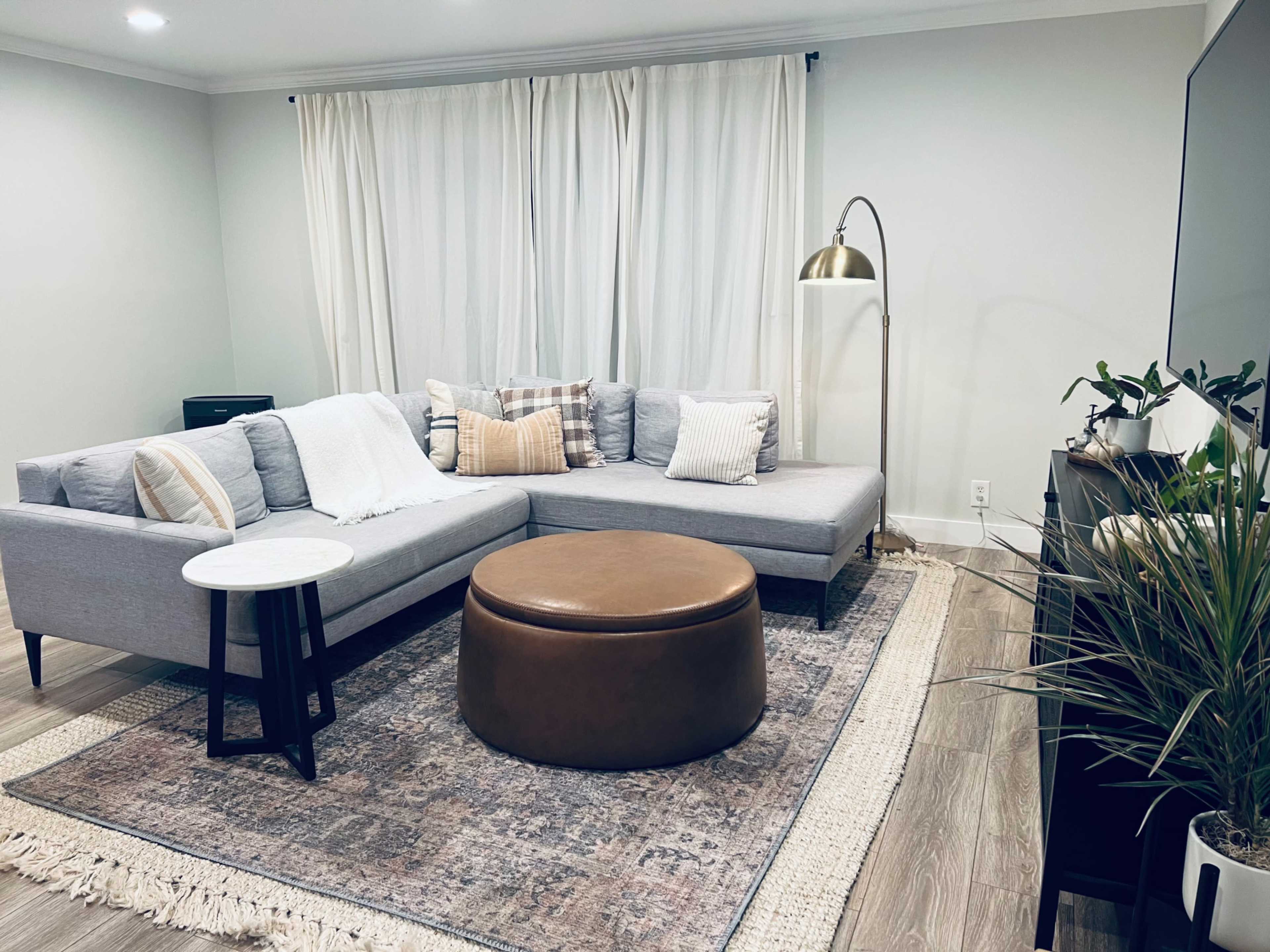 A gray sectional sofa with various cushions is positioned in a bright living room, accompanied by a round leather ottoman and a white side table on a patterned area rug.