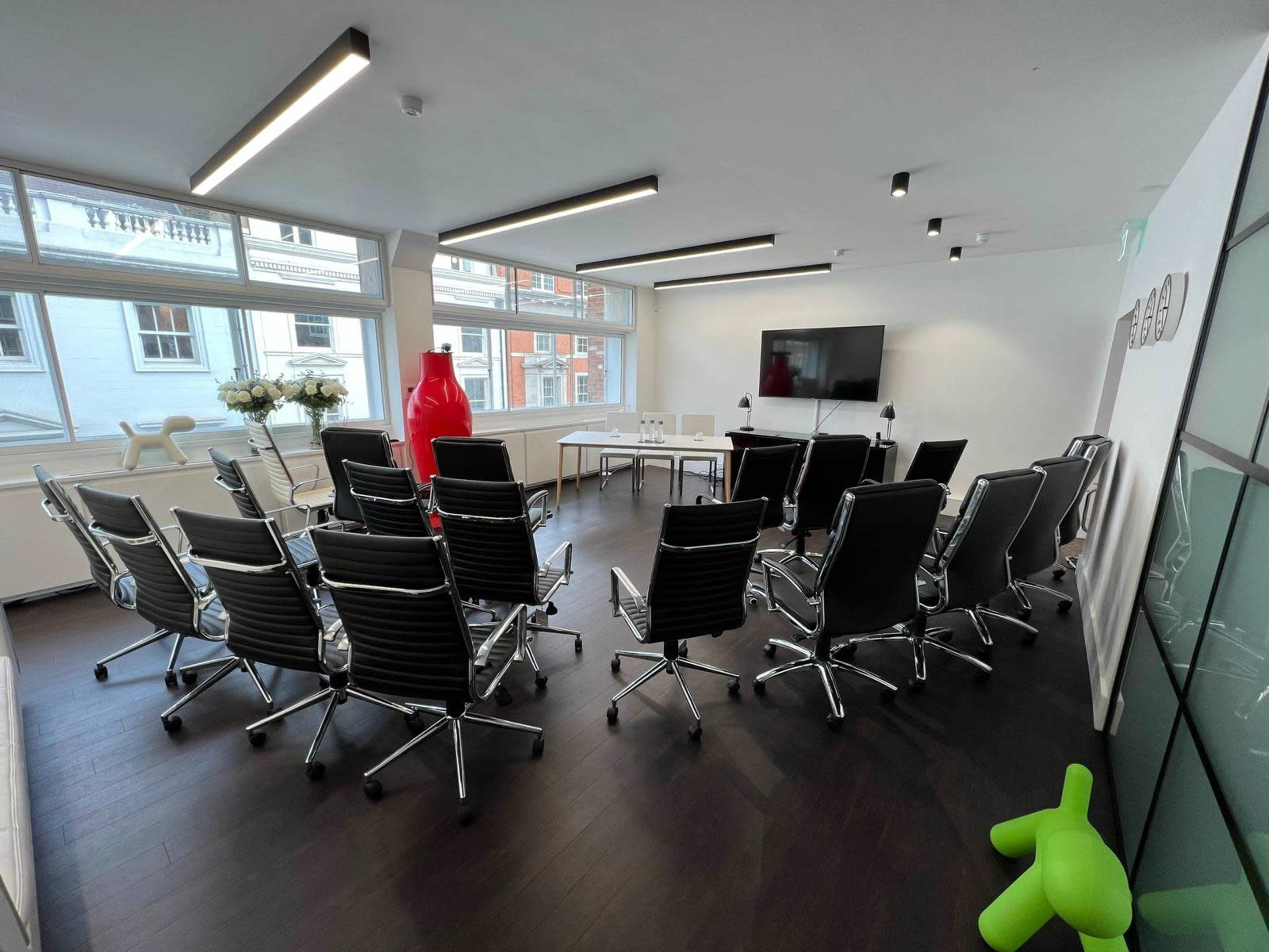 A modern conference room arranged with black rolling chairs facing a large television screen, with a table at the front and a green chair in the shape of a dog.