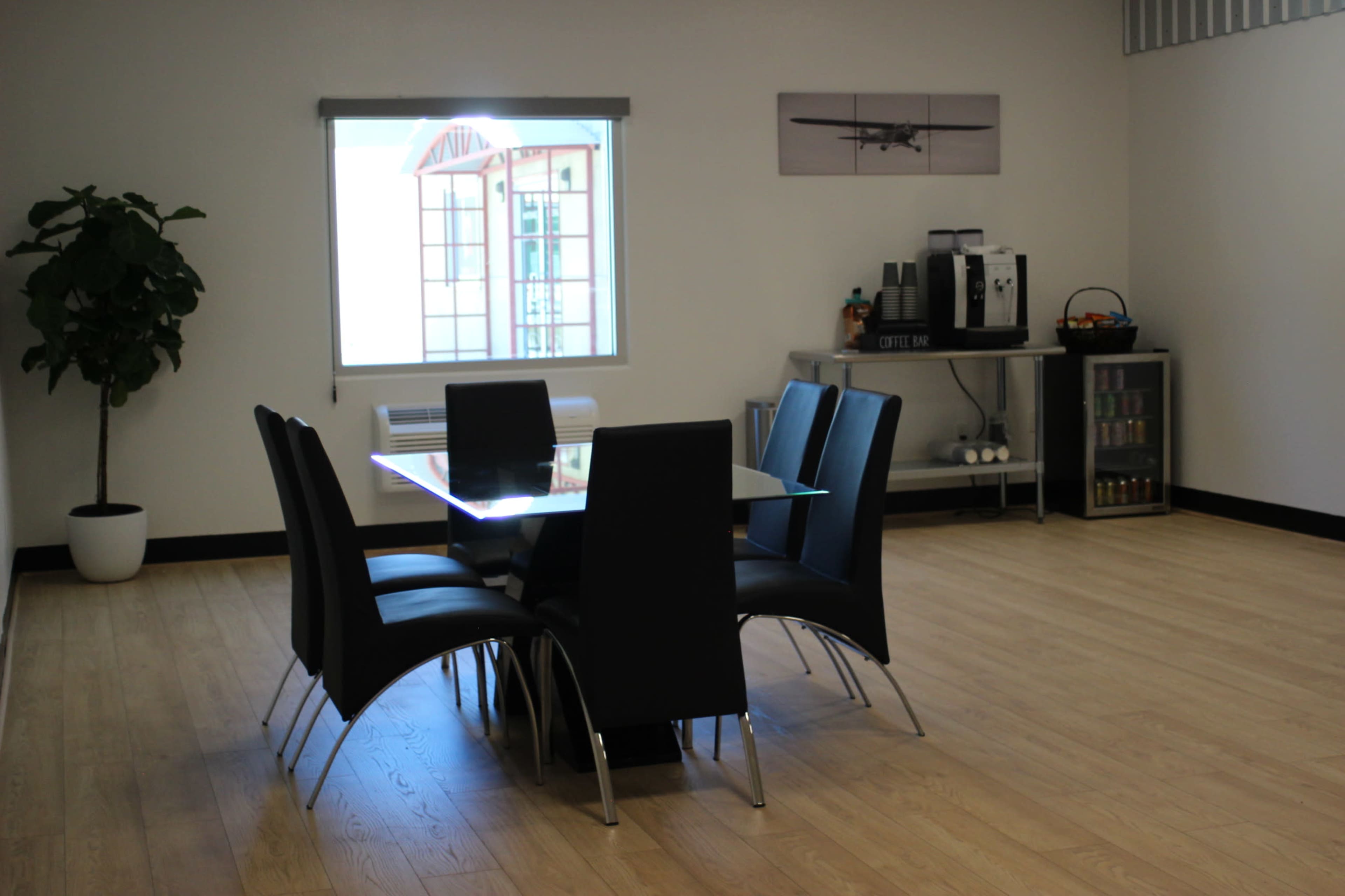 A dining area features a glass table surrounded by six black chairs, with a window and a coffee station in the background.