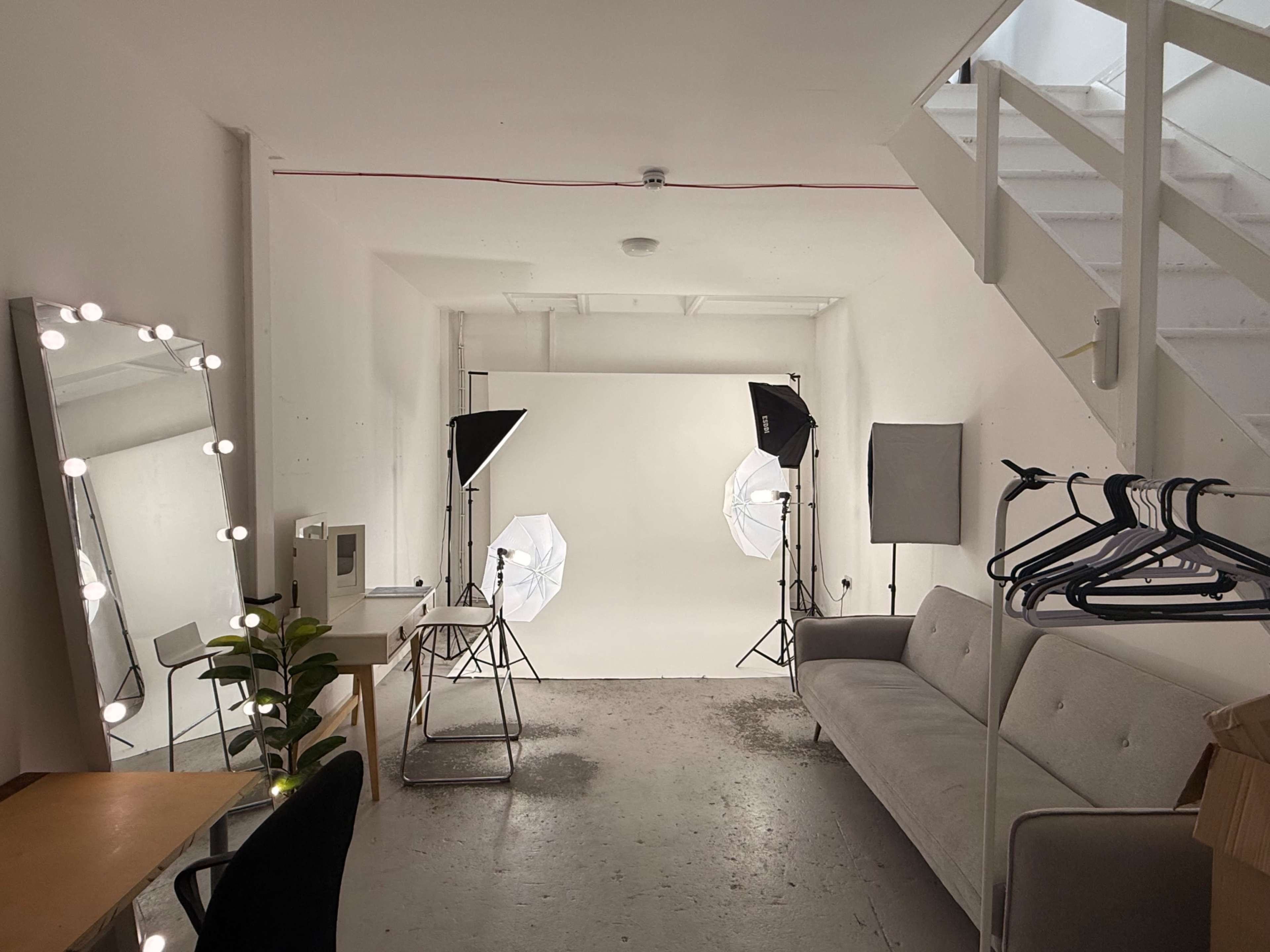The image shows a well-lit studio space with a gray sofa, a makeup mirror with lights, a desk, and several photography lights set up against a blank backdrop.