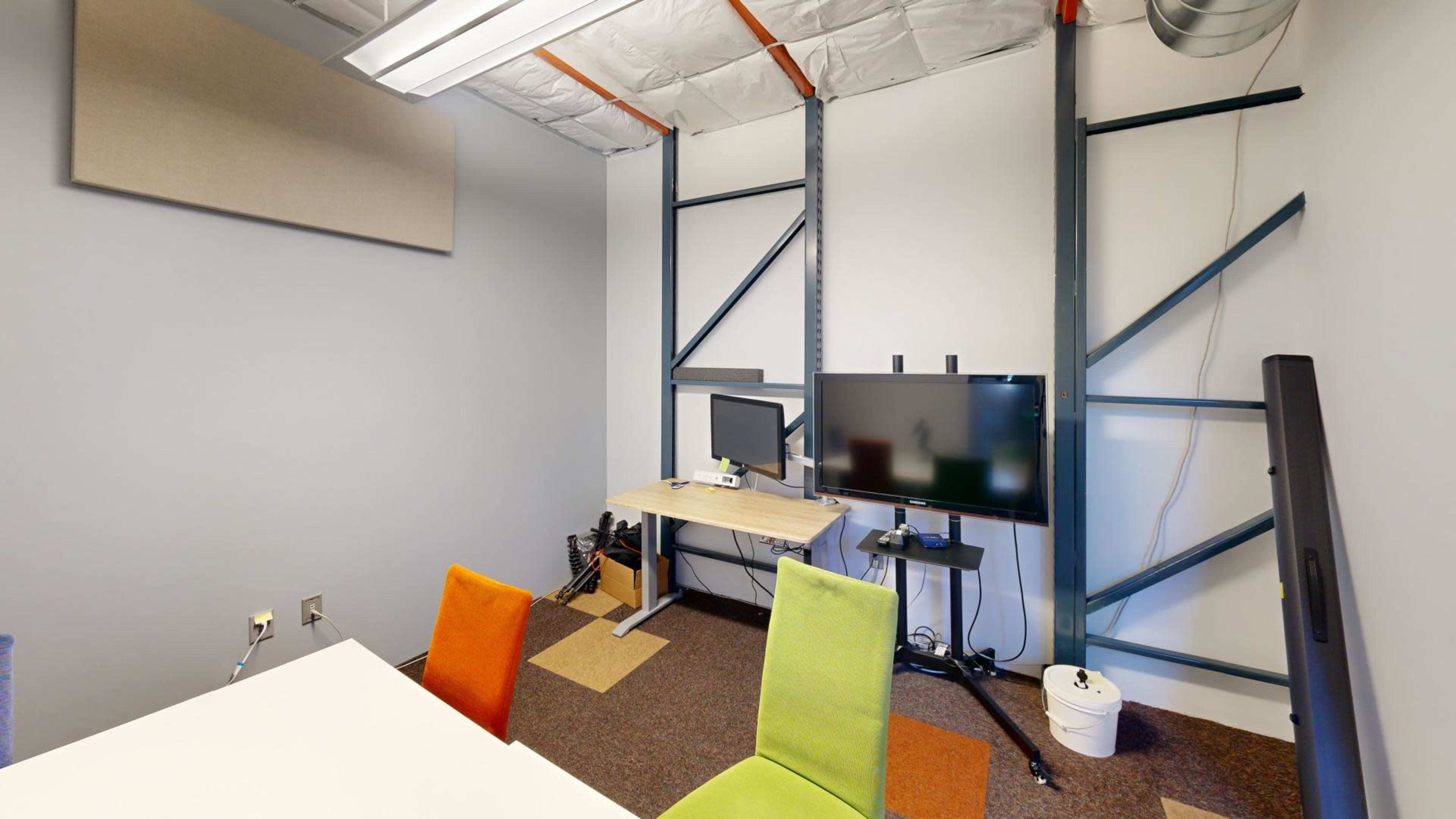 The image shows a small office space featuring a desk with two monitors, colorful chairs, and exposed structural elements on the walls.