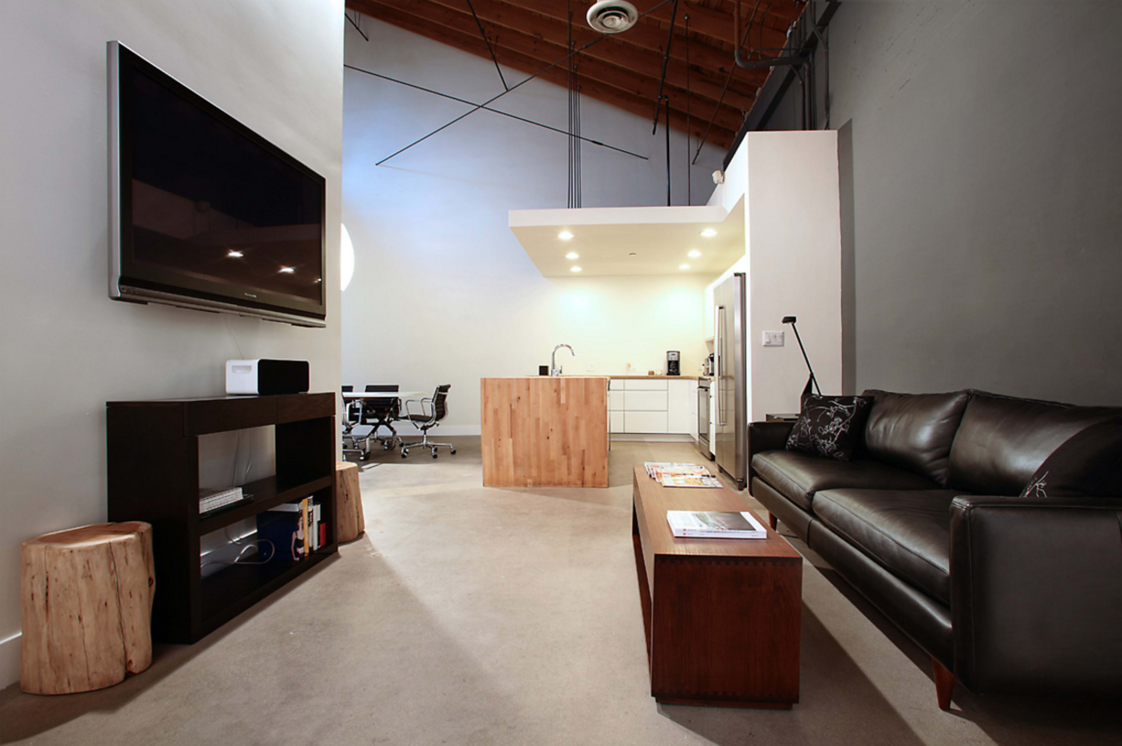 The image shows a modern open-concept living space featuring a black leather sofa, a wooden coffee table, a television mounted on the wall, and a kitchen area with a minimalist design.