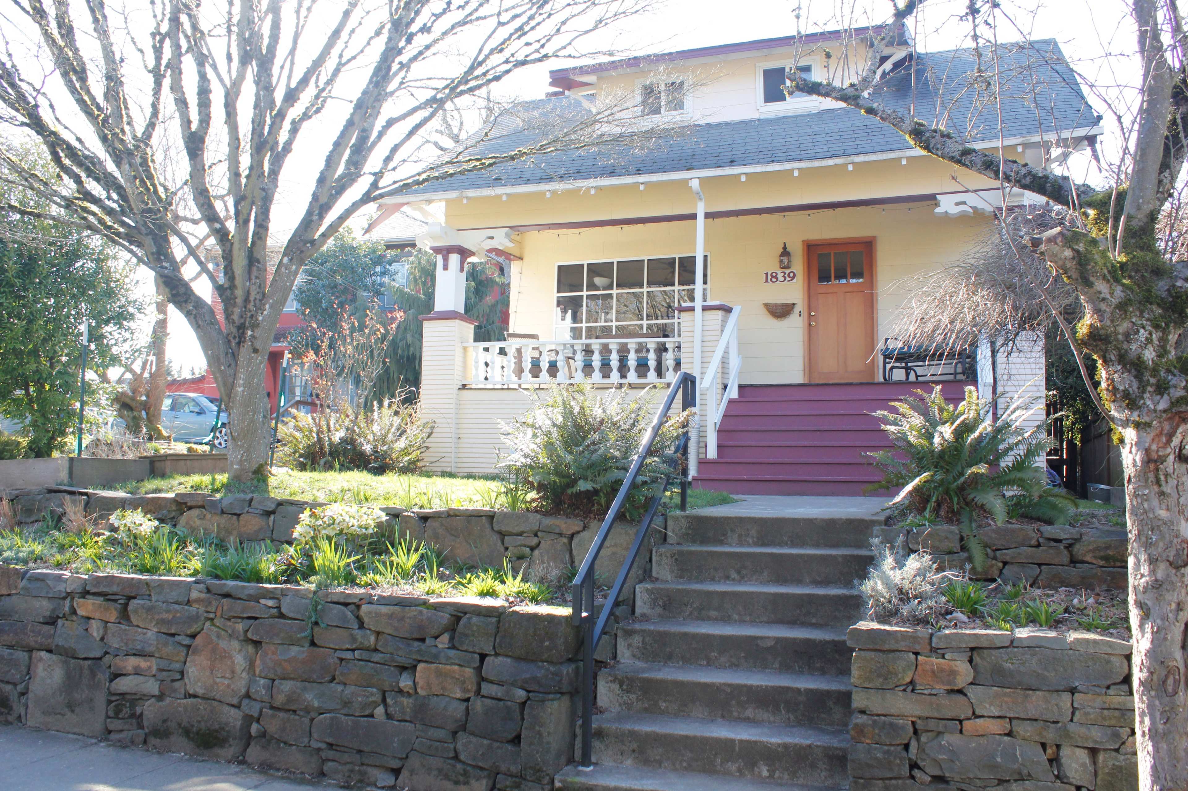 A two-story yellow house with a porch and stone steps is surrounded by trees and shrubs.