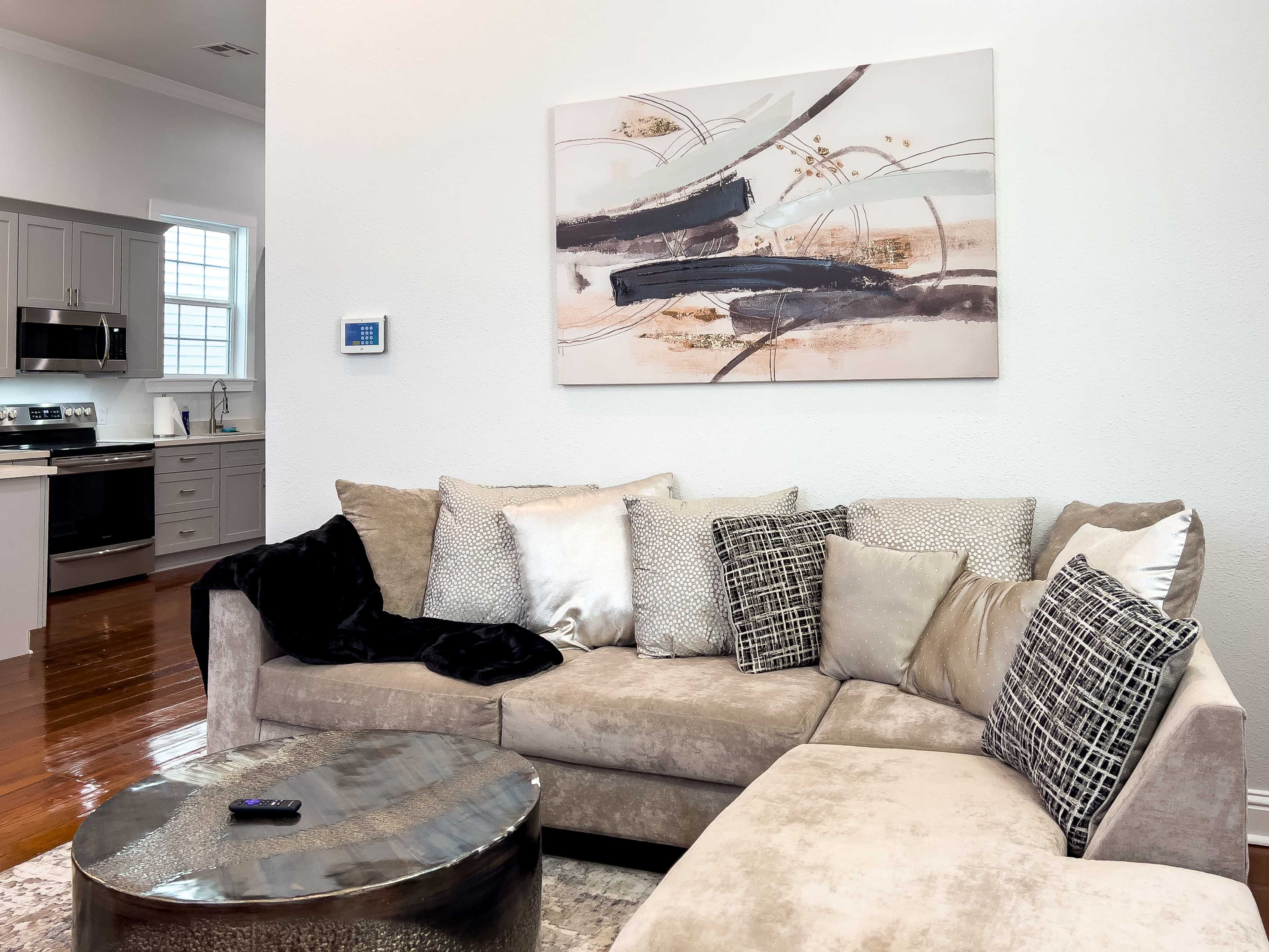 A modern living room features a sectional sofa with various pillows, a round metallic coffee table, and abstract wall art, with a glimpse of a kitchen in the background.