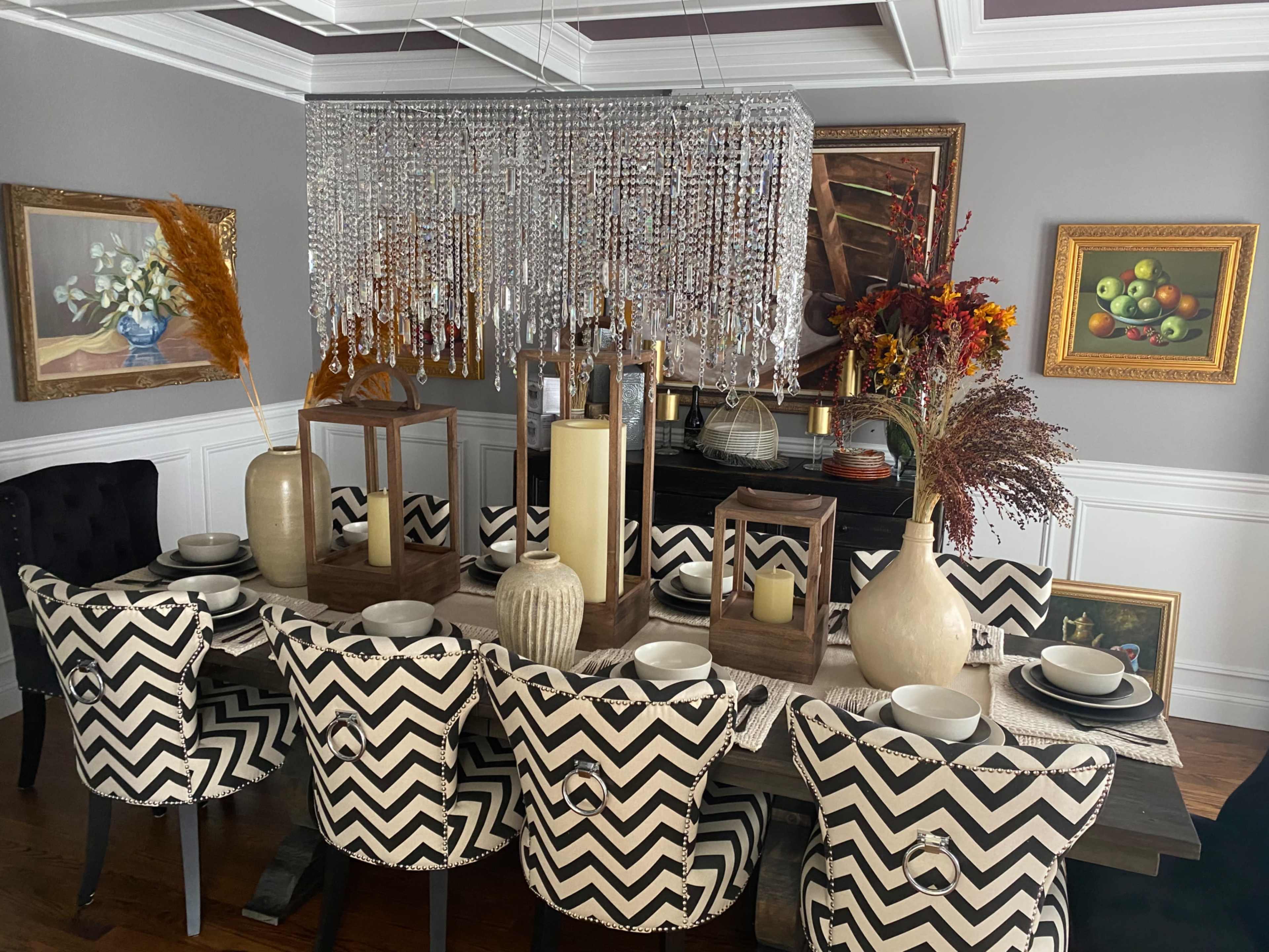 A dining room features a long table set with dishes and decorated with vases, surrounded by upholstered chairs with a zigzag pattern, and a crystal chandelier overhead.
