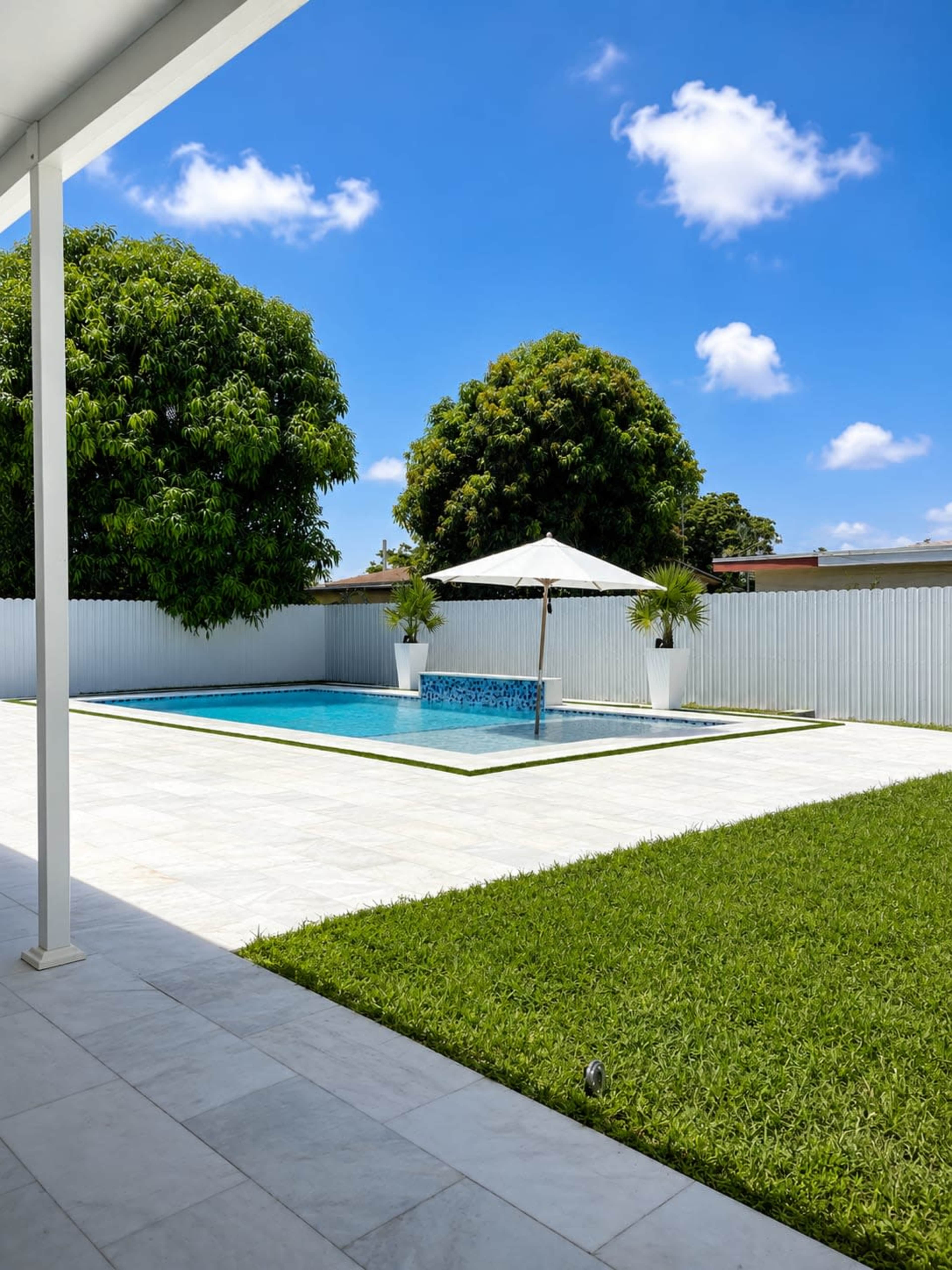 The image shows a backyard with a rectangular swimming pool surrounded by green grass, featuring a large umbrella over a seating area.