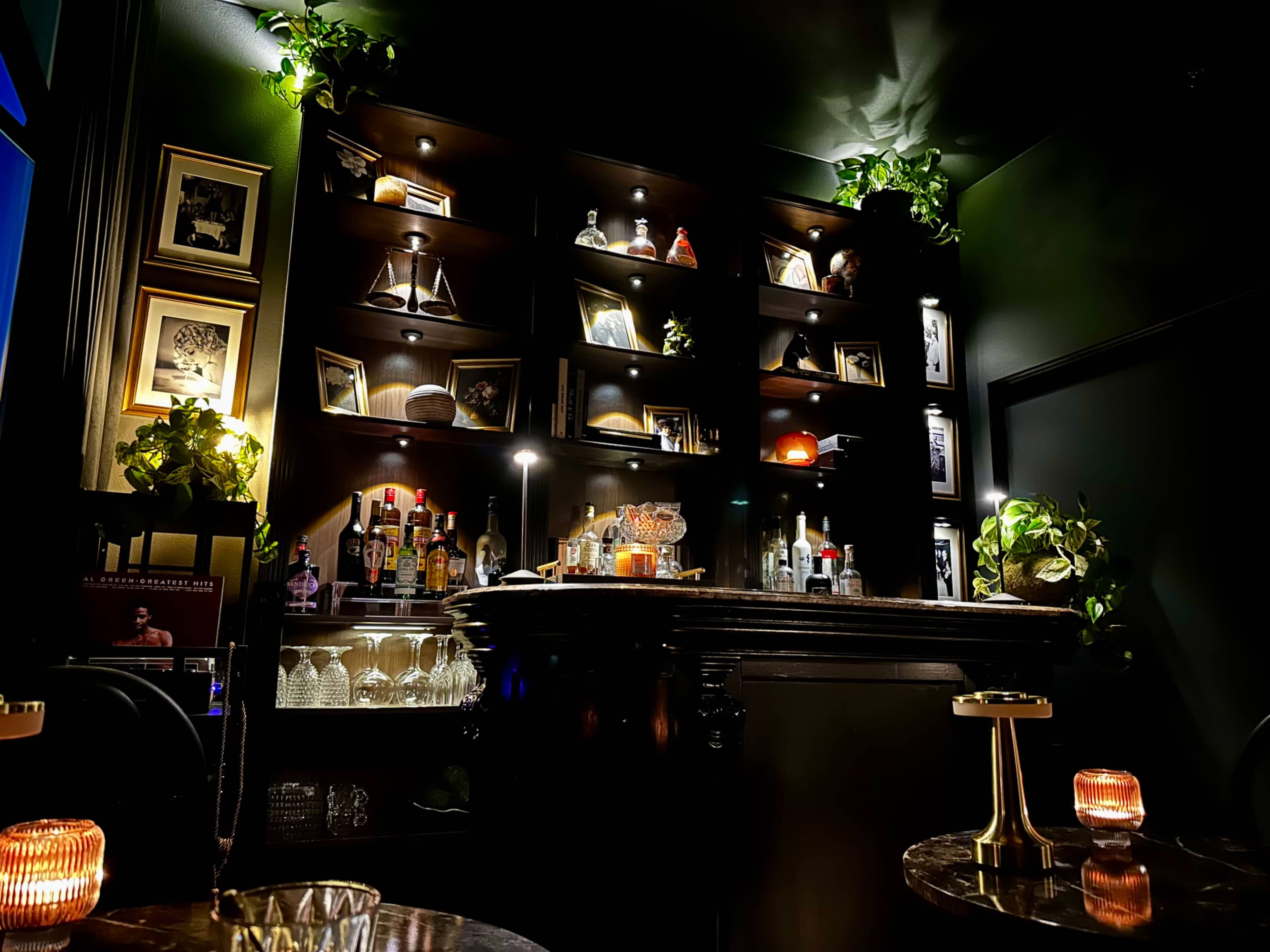 The image shows a dimly lit bar with shelves displaying various bottles and decorative items, along with a polished bar counter and small tables featuring candlelight.