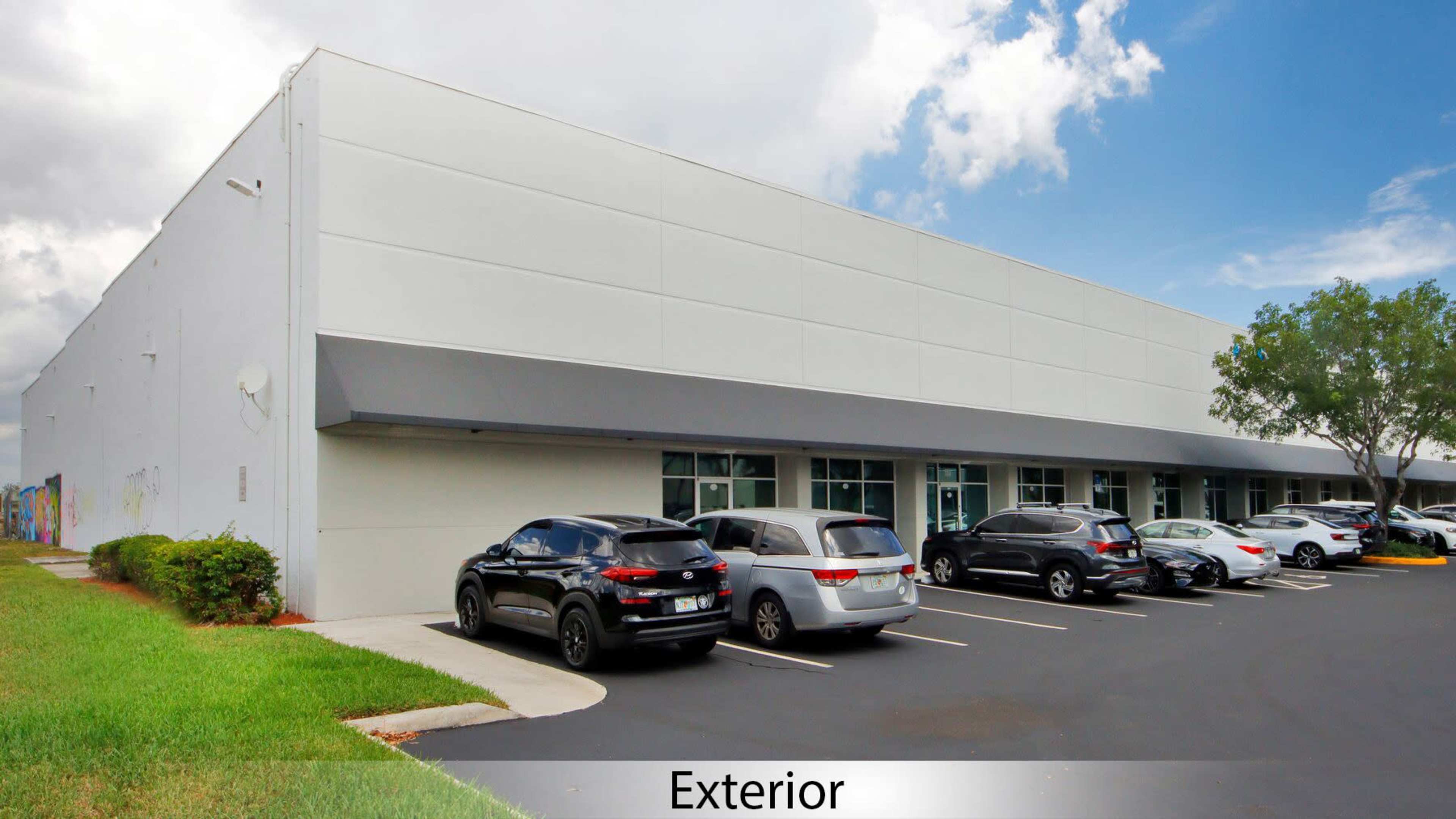 The image shows the exterior of a large, white commercial building with a parking lot filled with several vehicles.