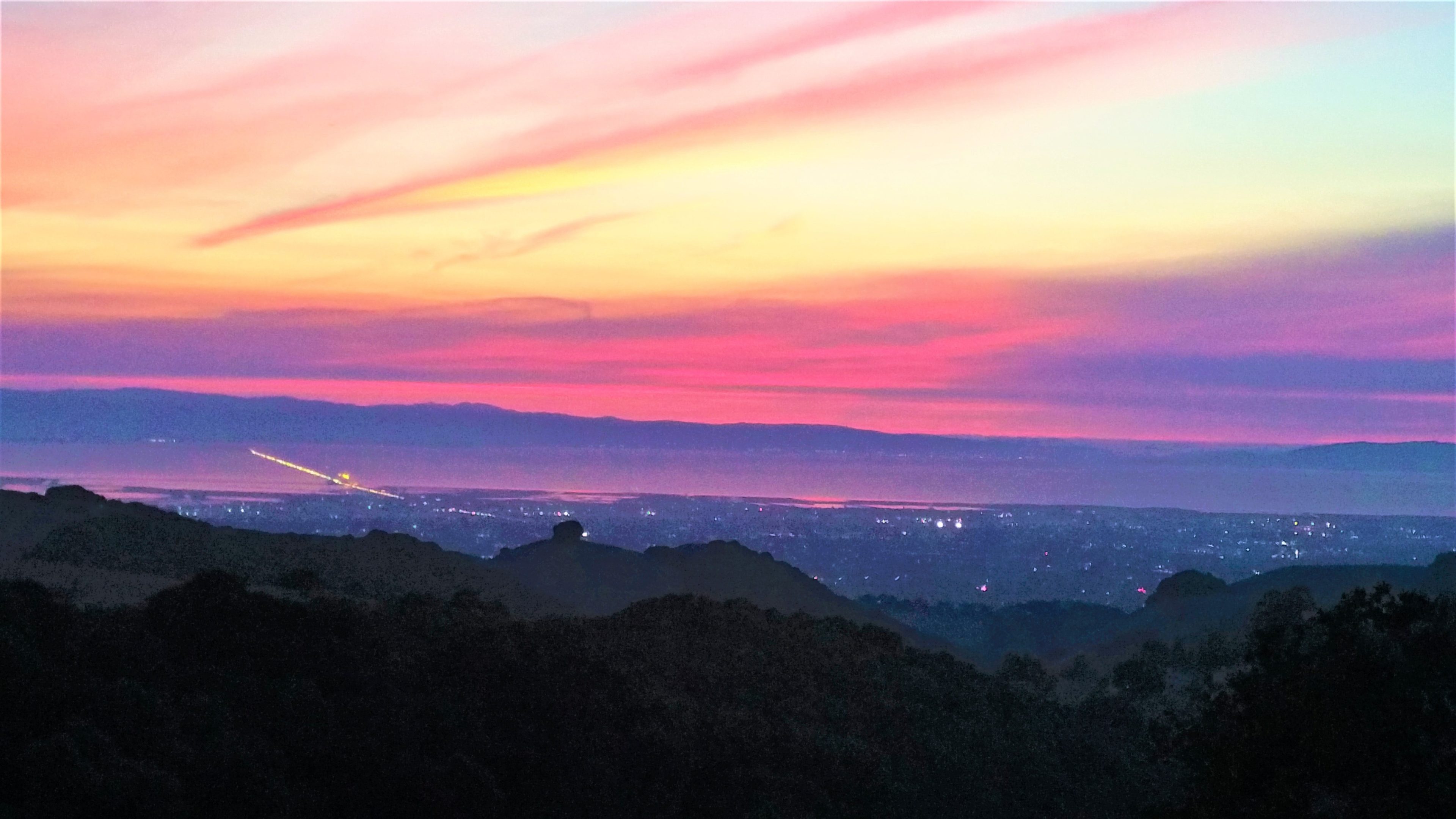 The image shows a panoramic view of a colorful sunset over a city and bay, with distant mountains and twinkling lights in the foreground.