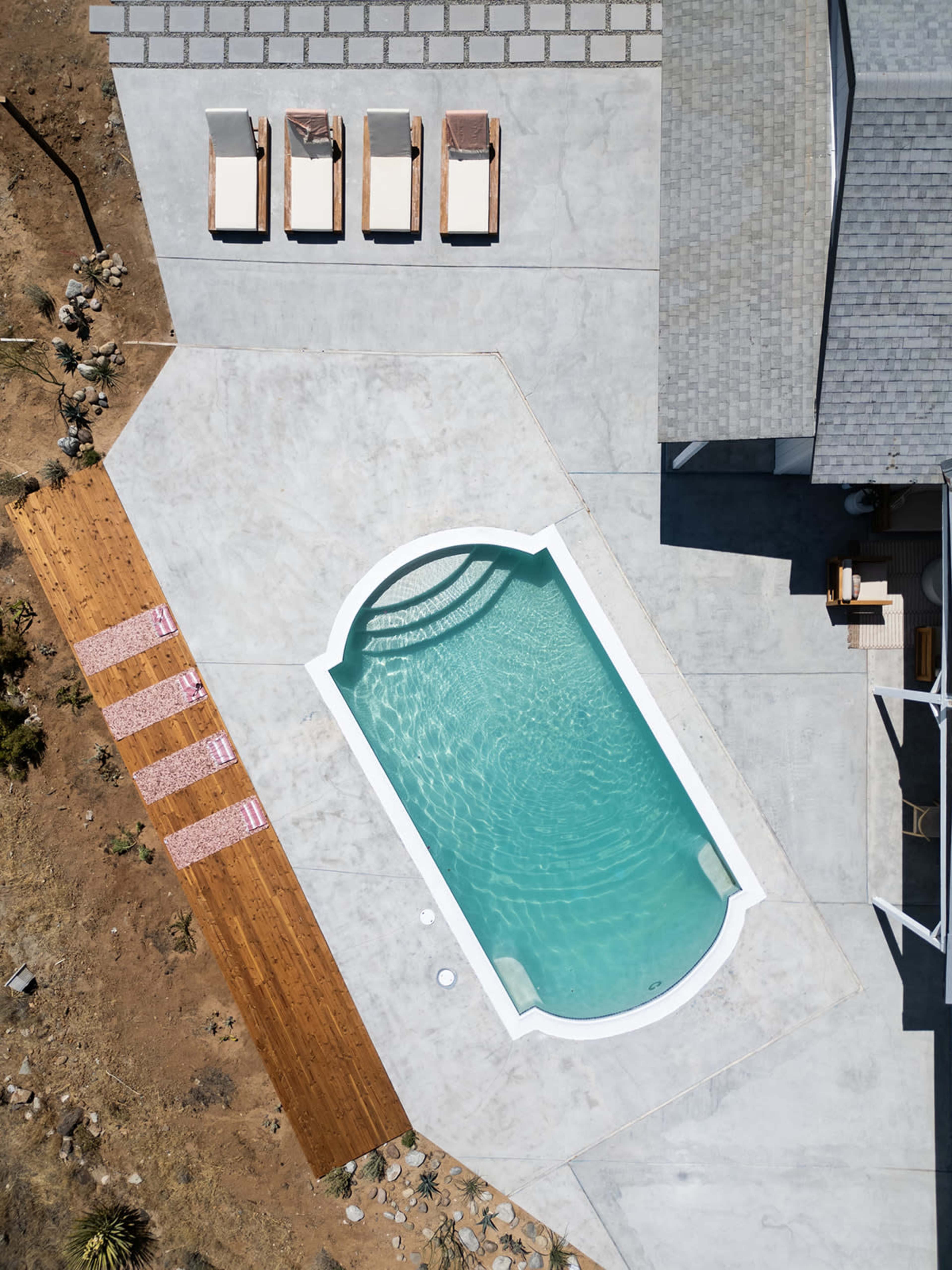 The image shows a rectangular pool surrounded by concrete and wooden decking, with lounge chairs arranged nearby and a house adjacent to the scene.