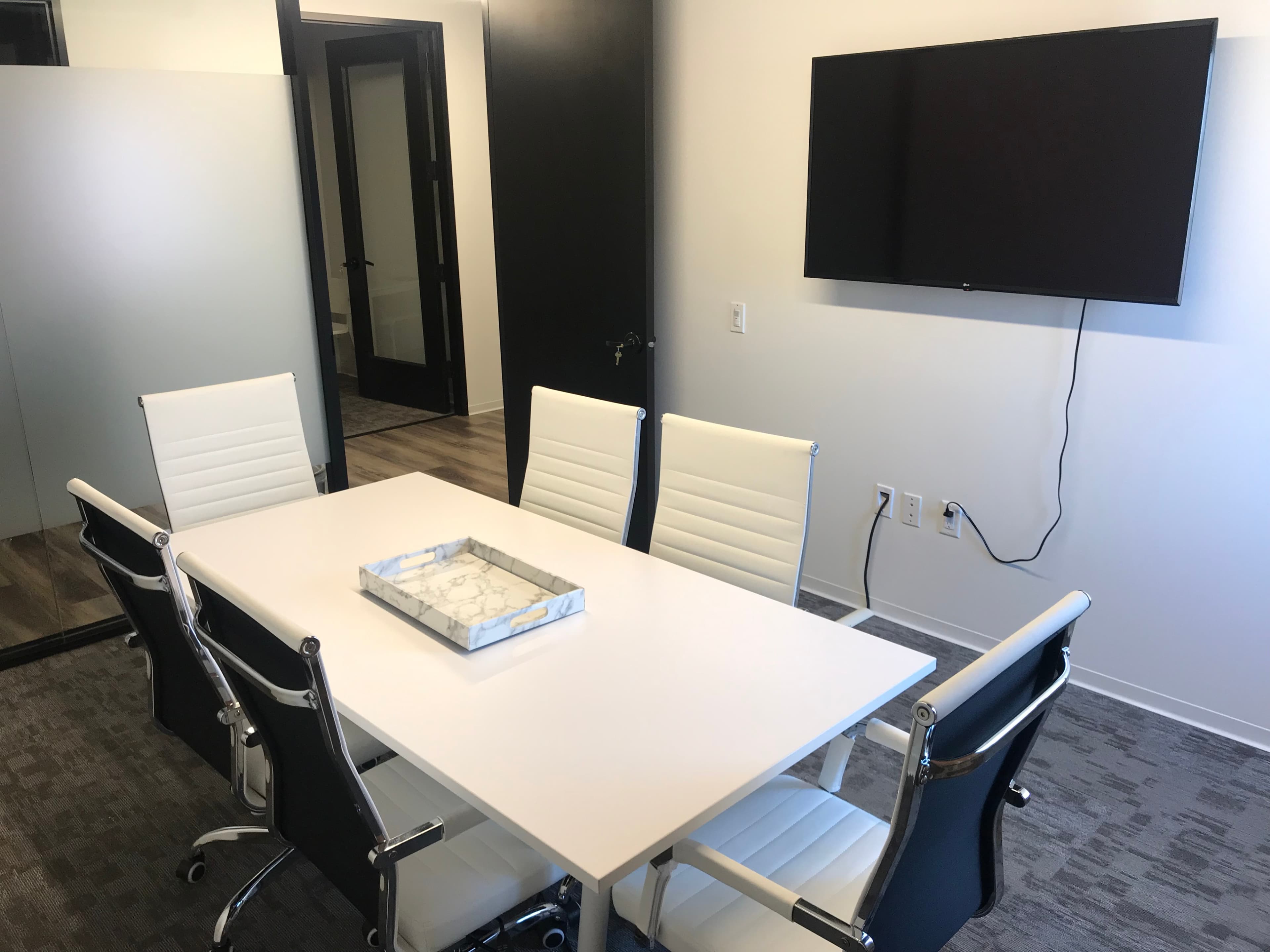 A modern conference room features a white table surrounded by six chairs and a wall-mounted television.