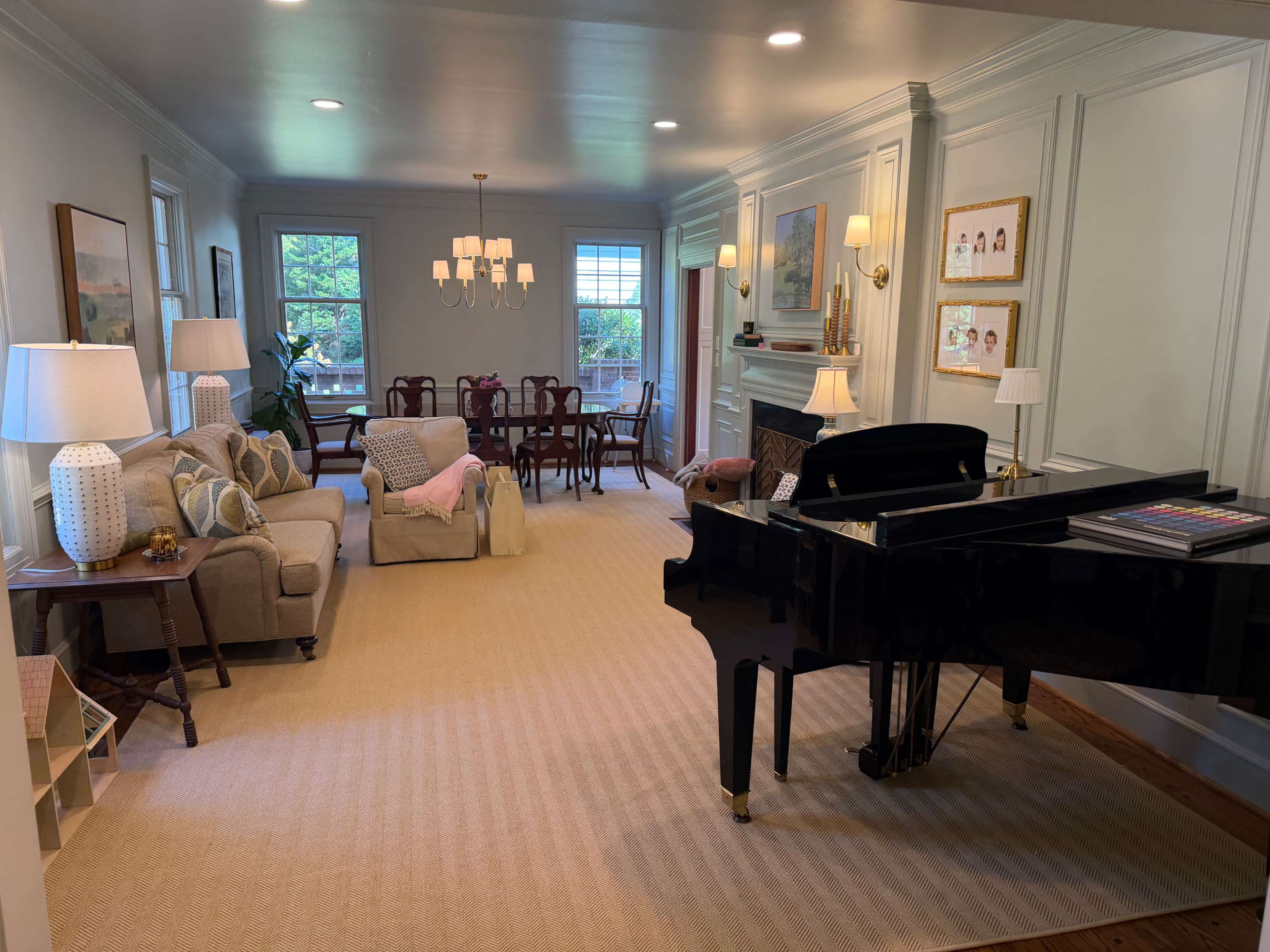 The image shows a spacious living room featuring a black grand piano, a beige sofa with decorative pillows, and a dining area with a wooden table and chairs.