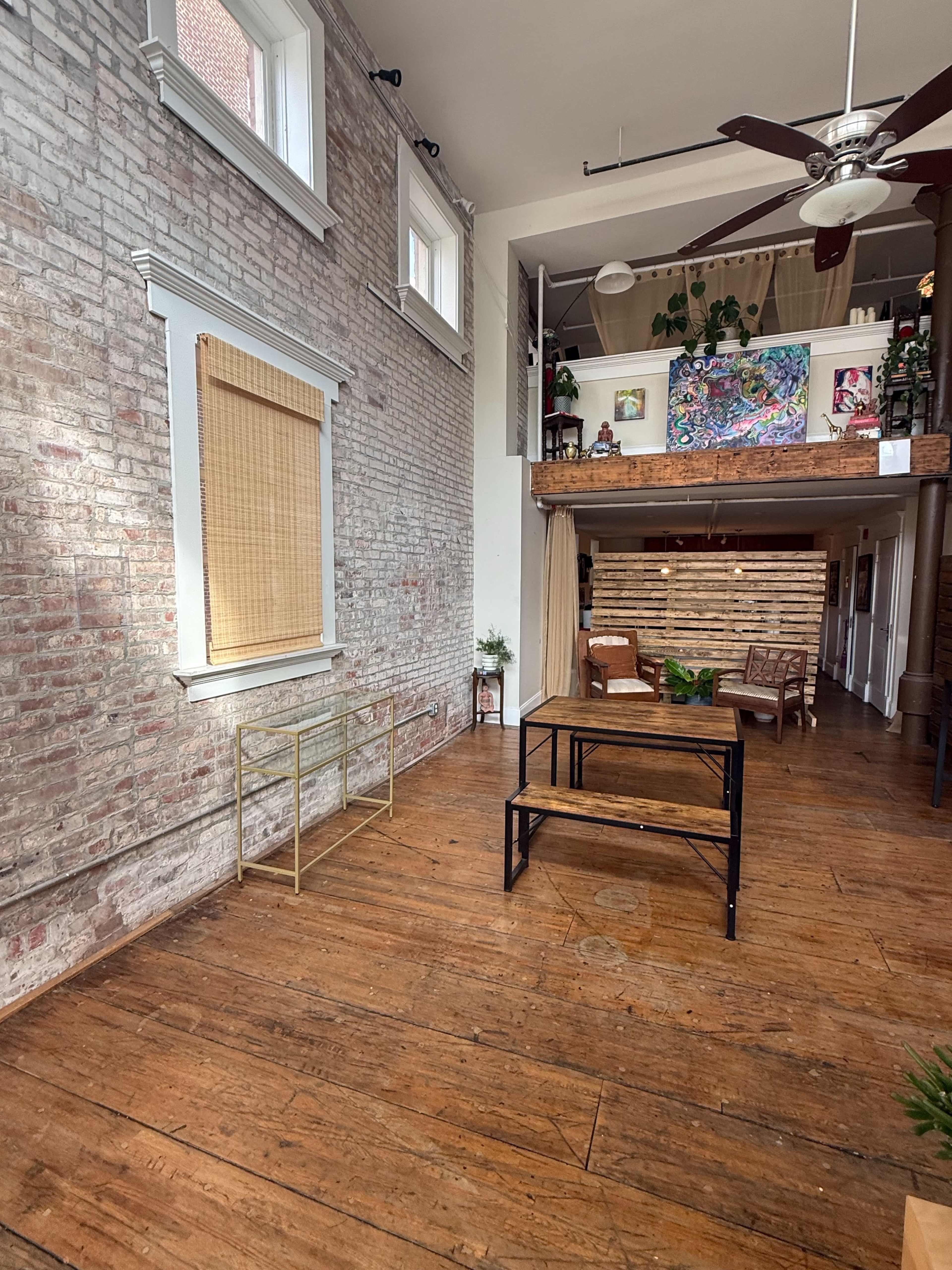 The image shows an interior space with exposed brick walls, hardwood flooring, a wooden table, and various plants, complemented by large windows and a high ceiling.