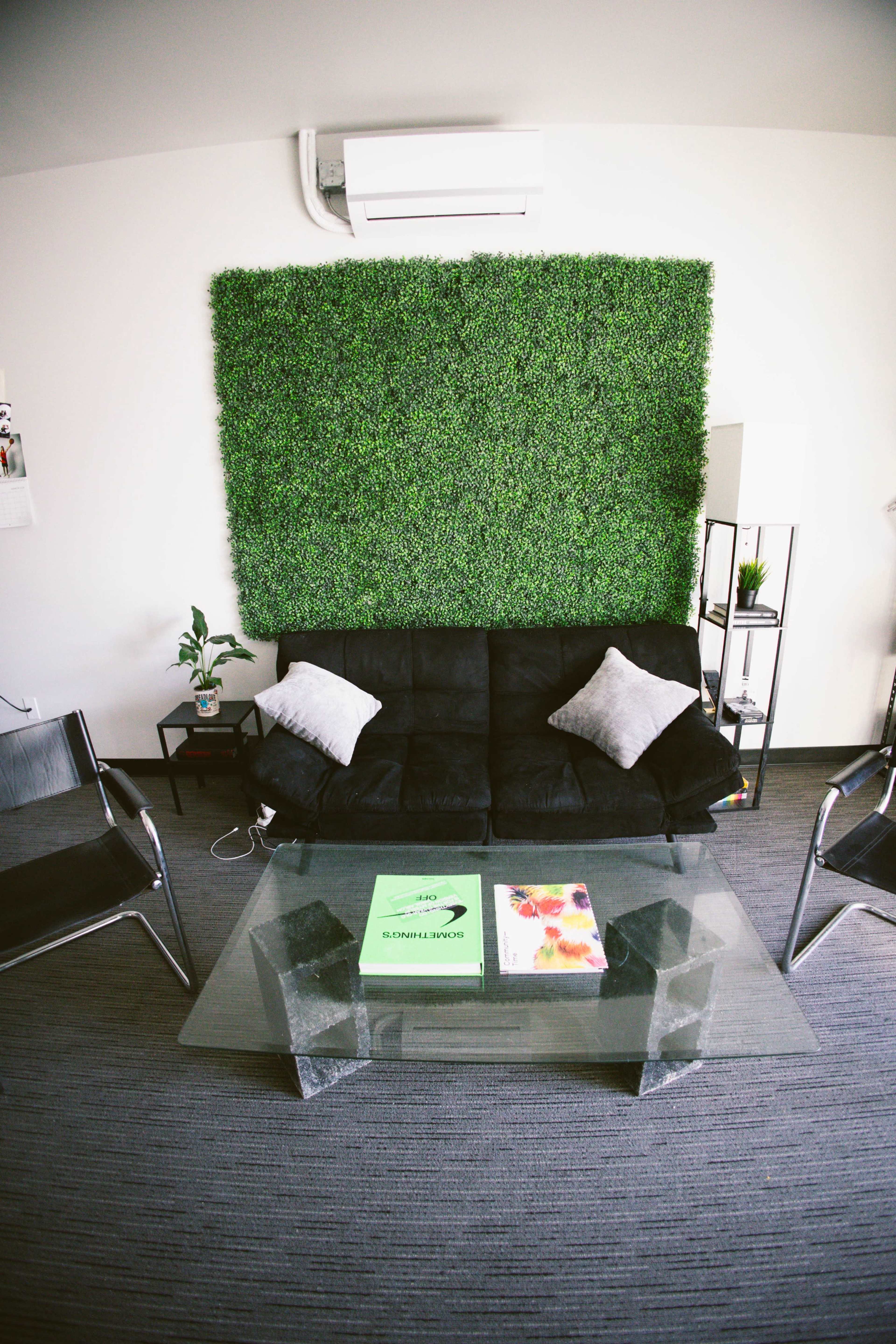 A modern living room with a black couch, a glass coffee table, and a green moss wall in the background, accompanied by indoor plants and two chrome chairs.