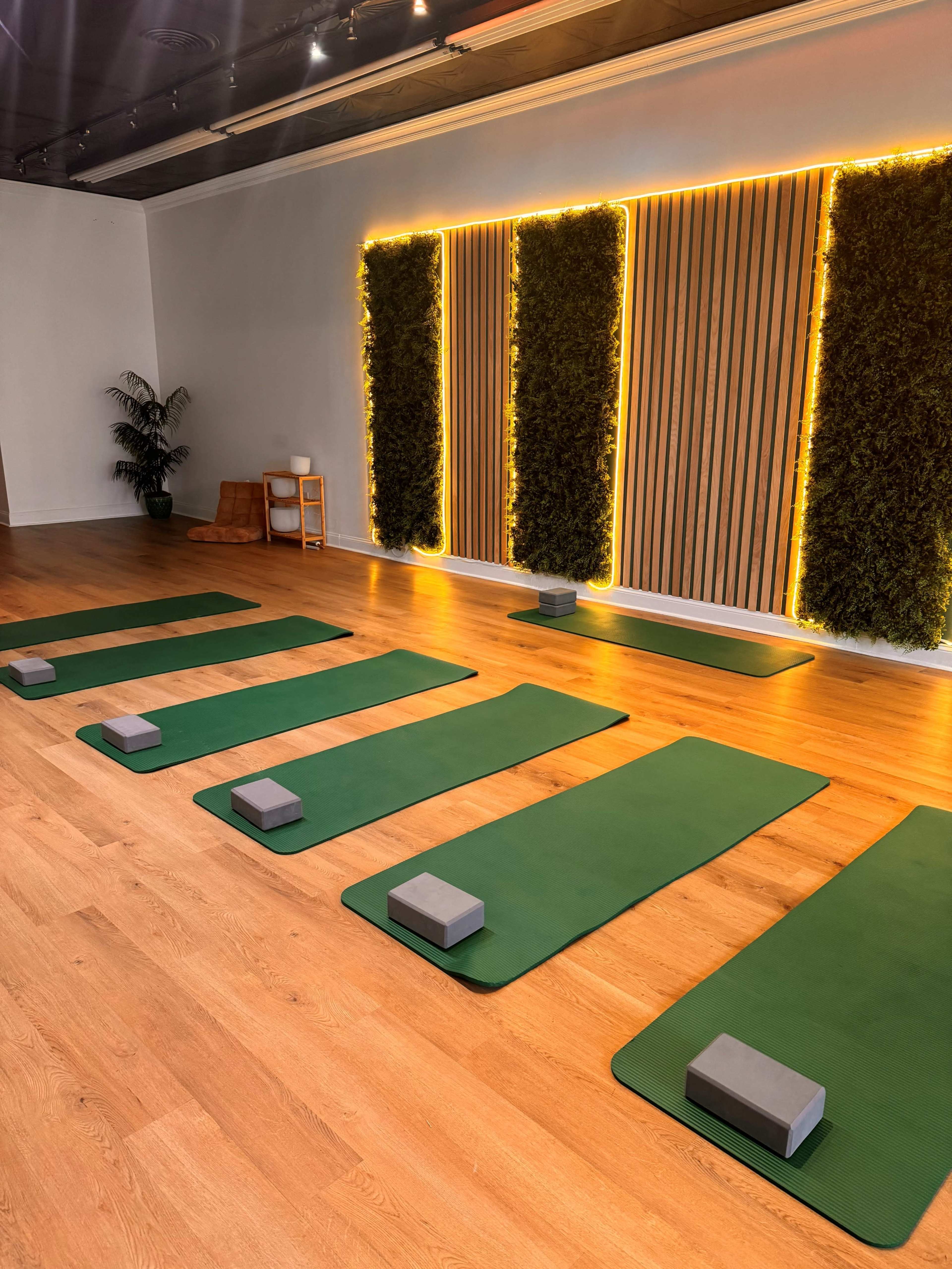 The image shows a yoga studio with several green mats and gray blocks arranged neatly on a wooden floor near a wall featuring greenery and ambient lighting.