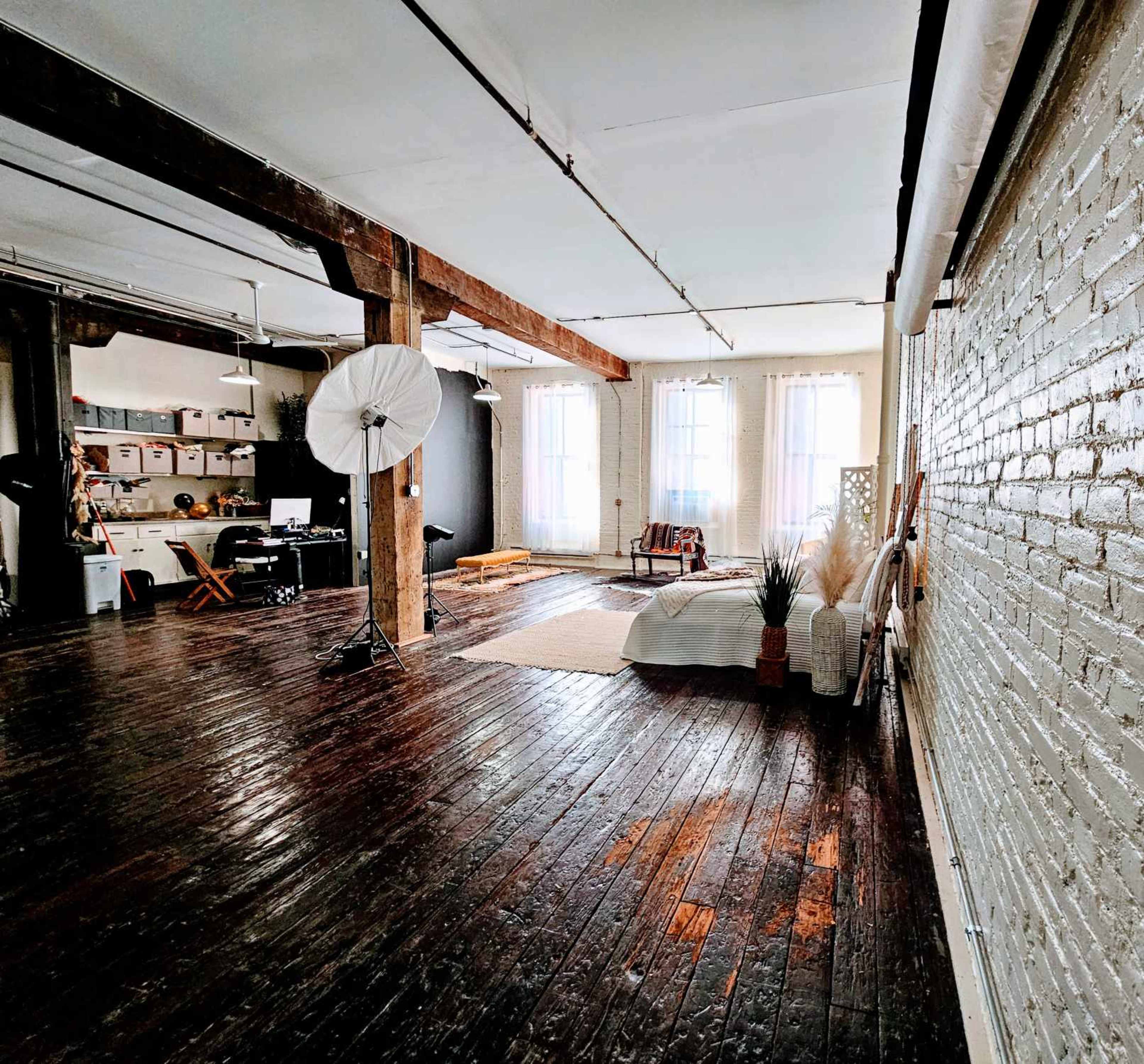 The image shows a spacious, well-lit studio with wooden floors, exposed beams, and a sectional area featuring a bed, plants, and a photography umbrella.