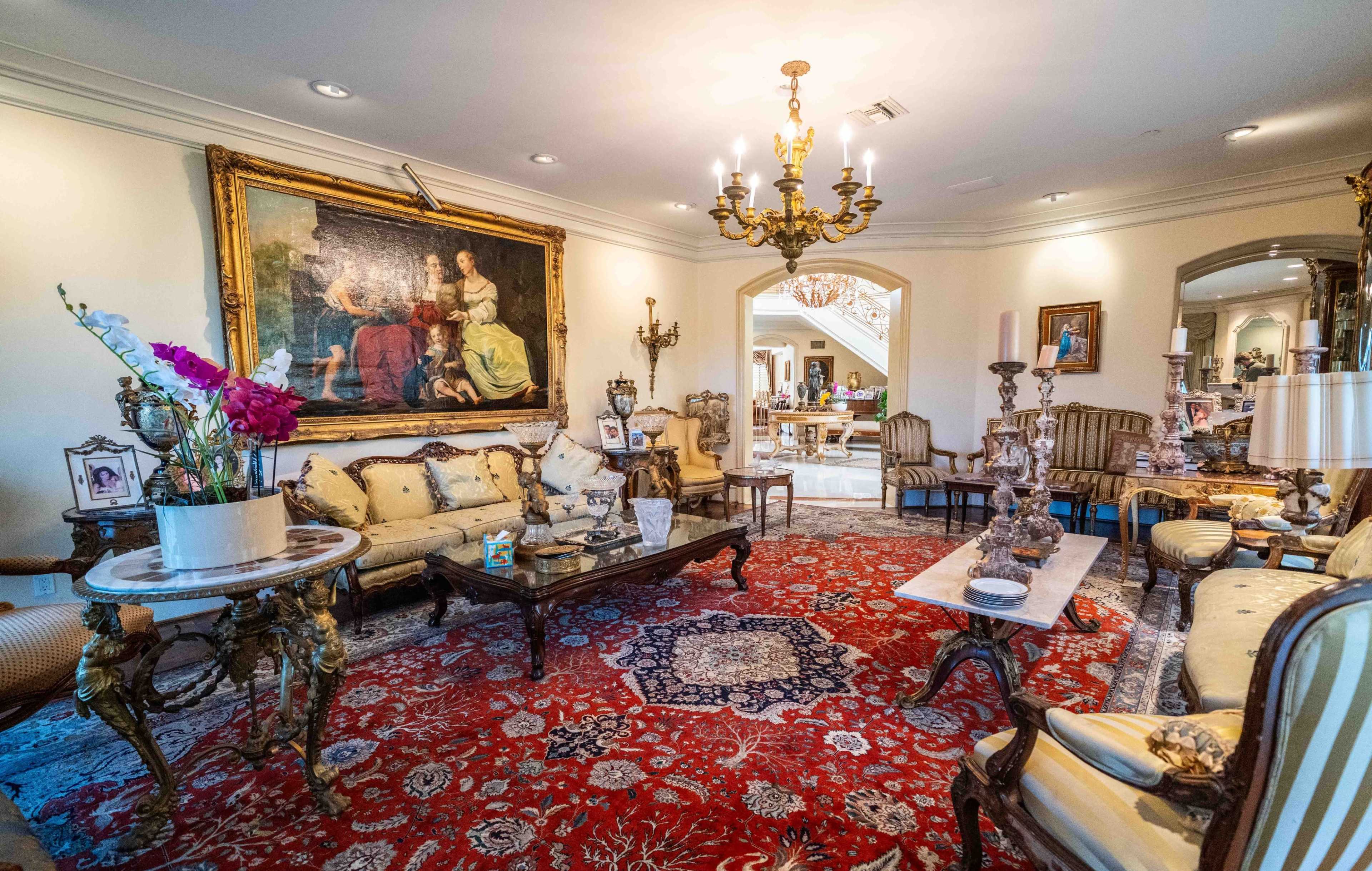 The image shows a lavishly decorated living room featuring ornate furniture, a large painting on the wall, and a red patterned rug.