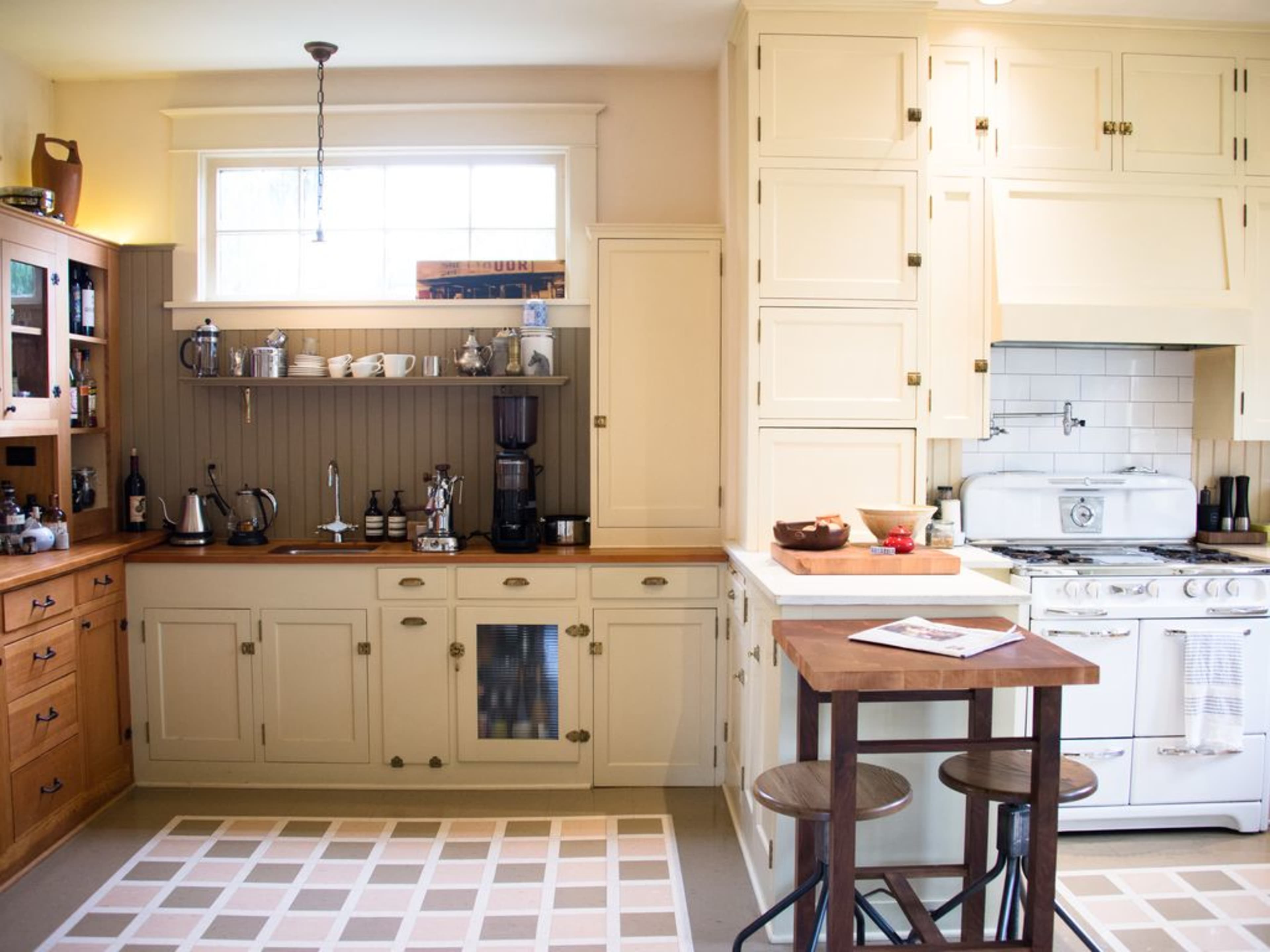 A modern kitchen featuring light-colored cabinets, a white stove, a coffee maker, and a small dining table with two stools against a backdrop of a patterned rug.
