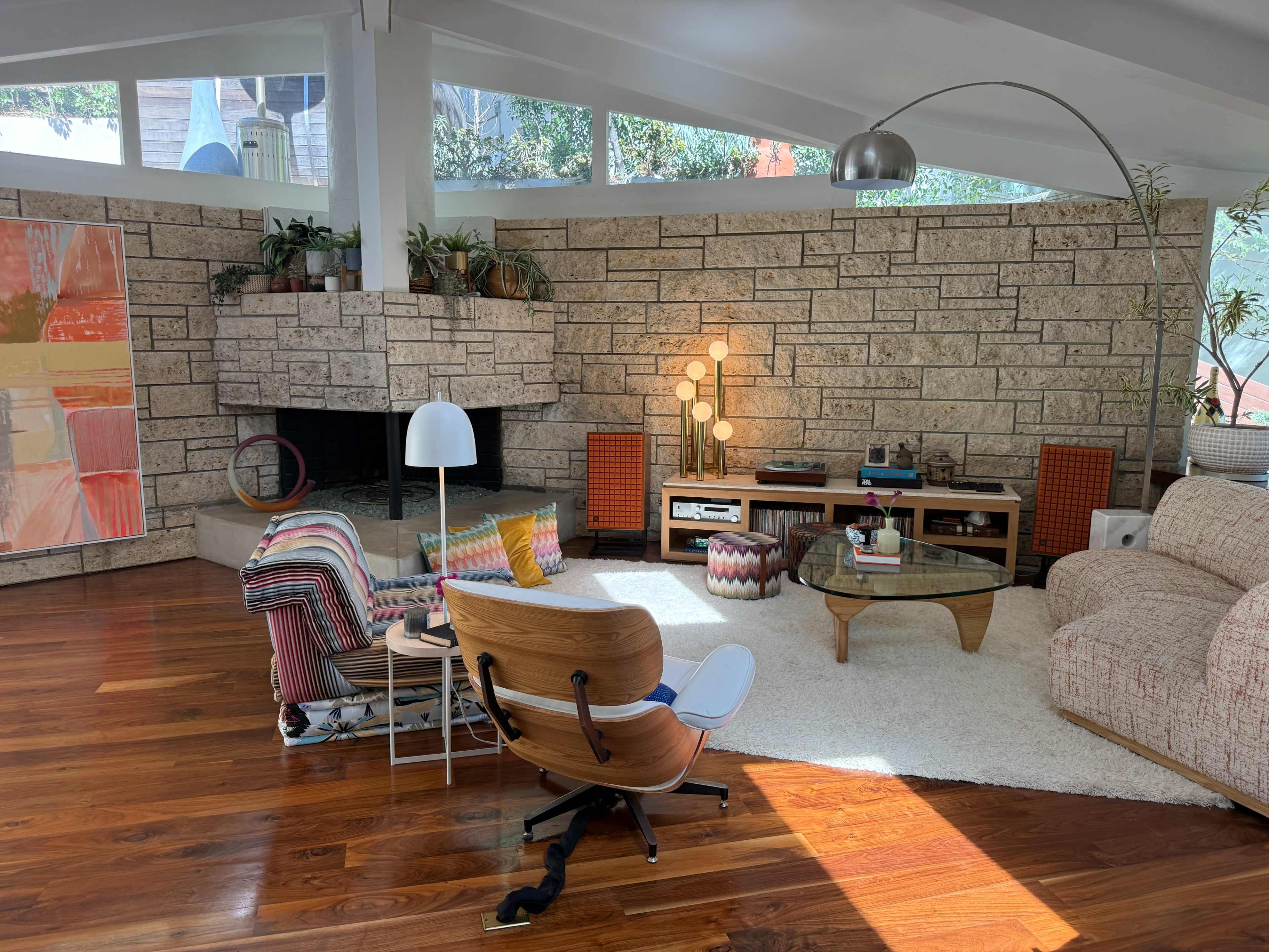 The image shows a mid-century modern living room featuring a stone fireplace, a large window allowing natural light, and various seating options arranged around a circular coffee table.