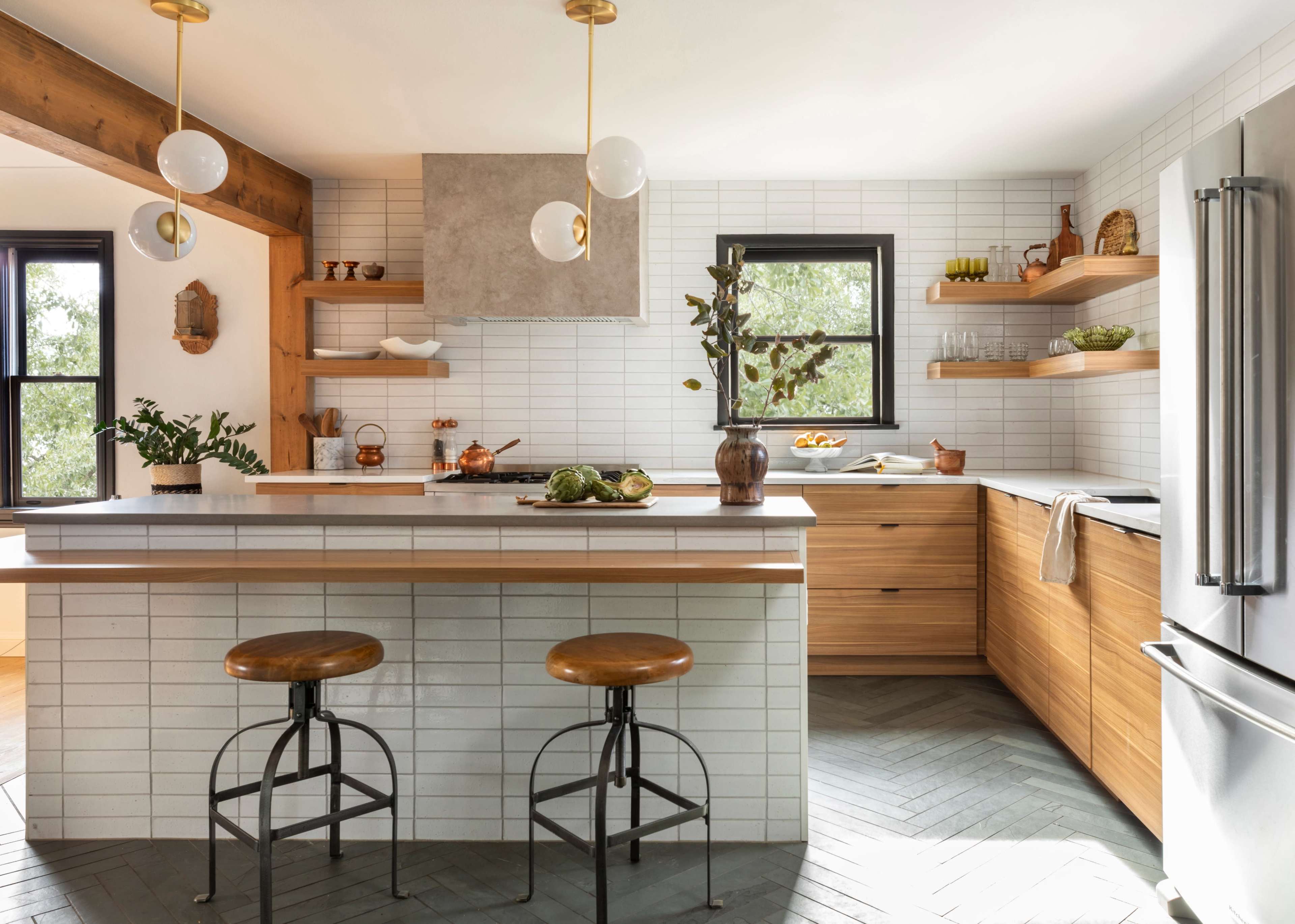 A modern kitchen features a wooden island with two stools, light-colored cabinetry, and open shelving filled with kitchenware.