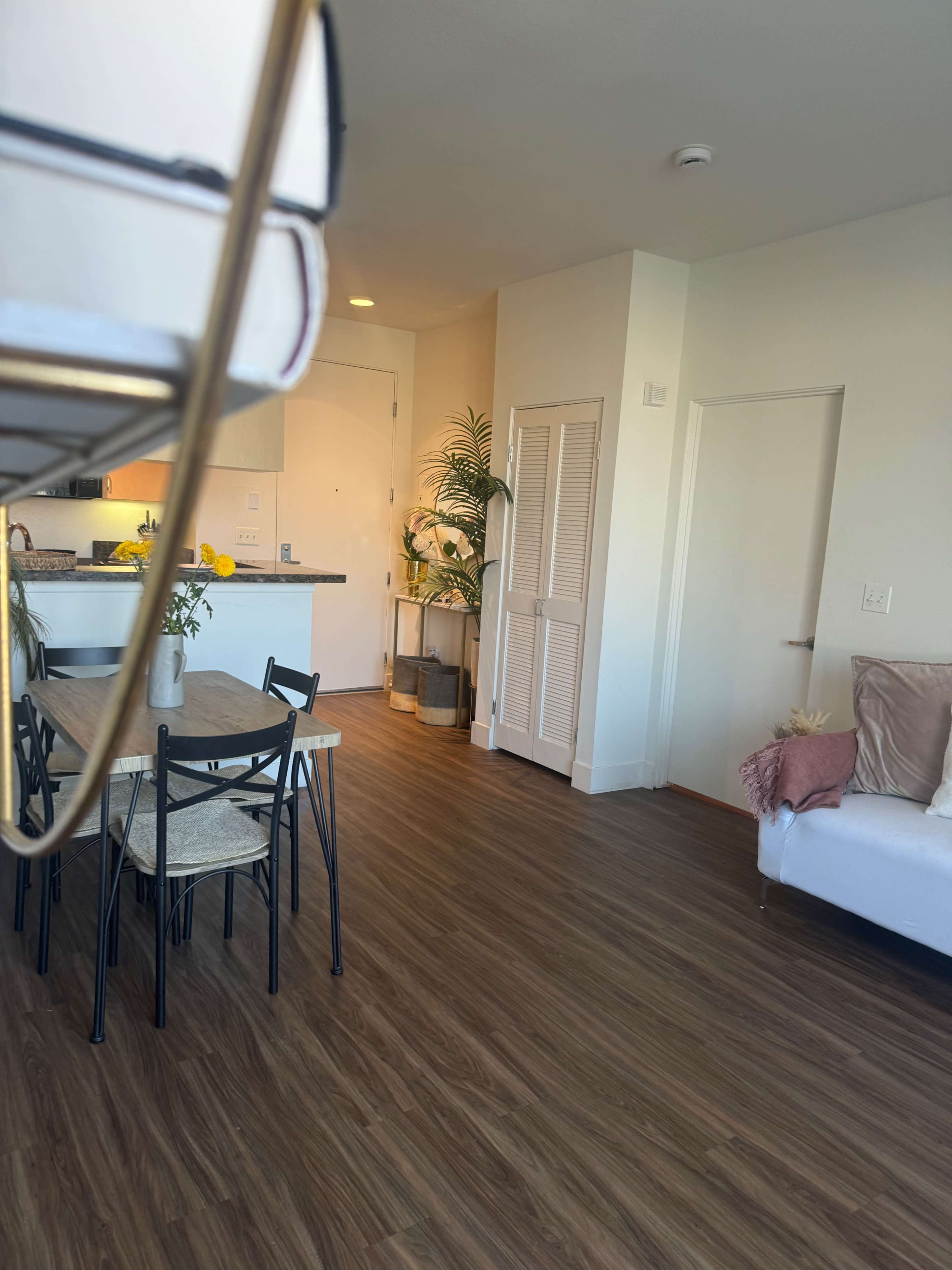 The image shows a modern living space featuring a dining table with chairs, a white sofa, and a kitchenette in the background.