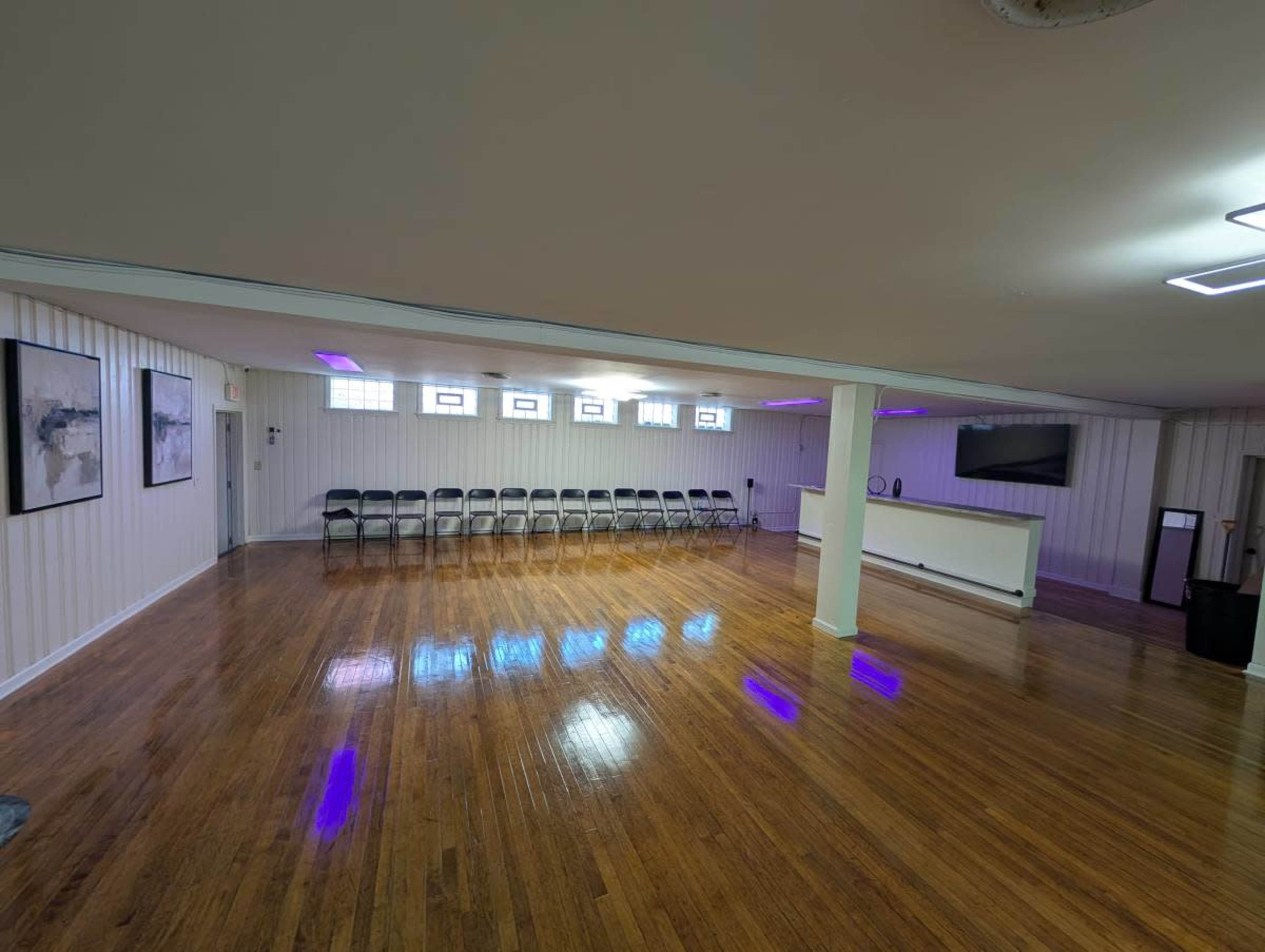 The image shows a spacious, empty room with polished wooden floors and rows of chairs lined up against one wall, illuminated by purple lighting.