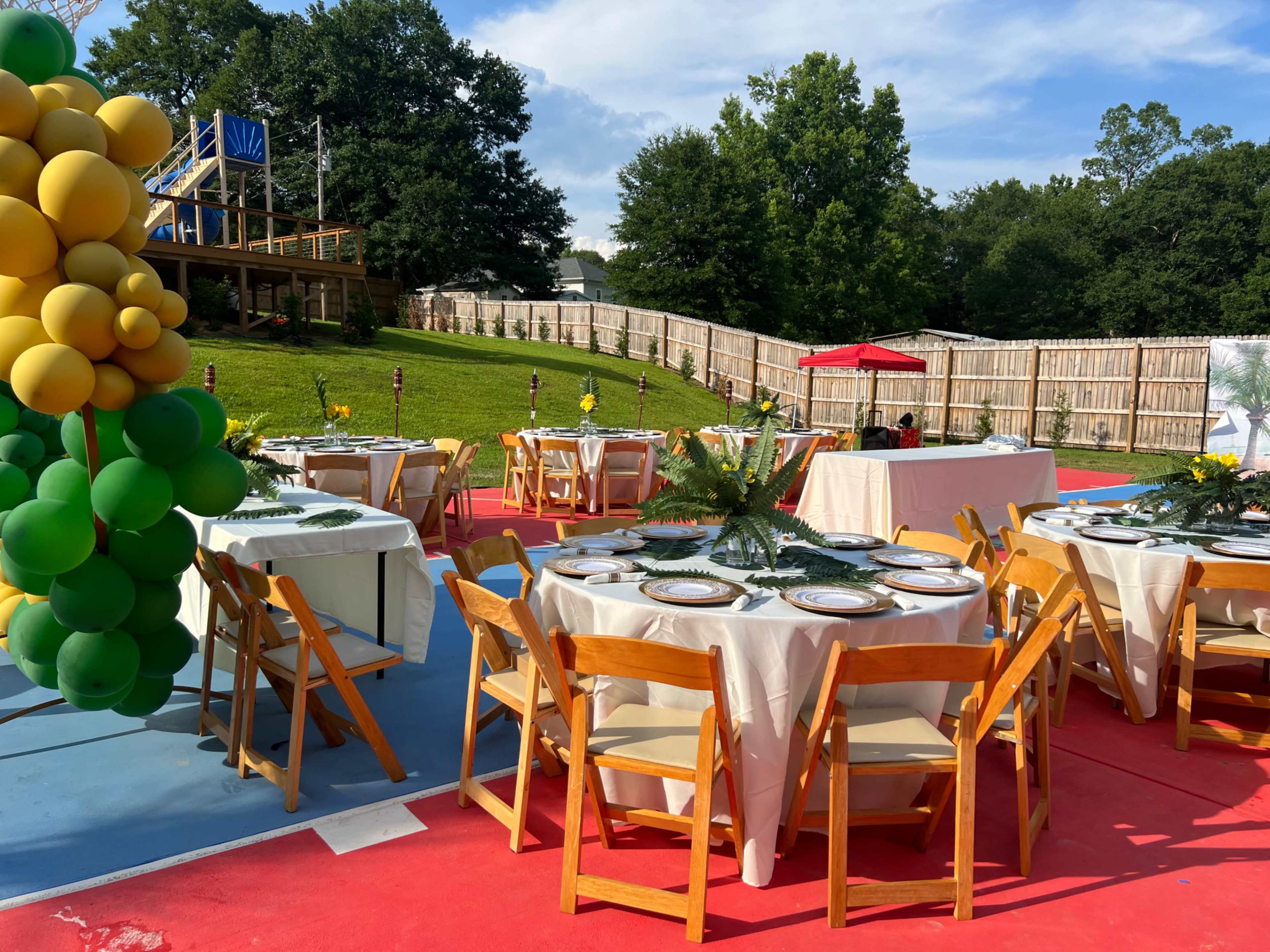 The scene shows a set-up for an outdoor event with several round tables adorned with centerpieces, surrounded by green grass and trees, and a play structure in the background.