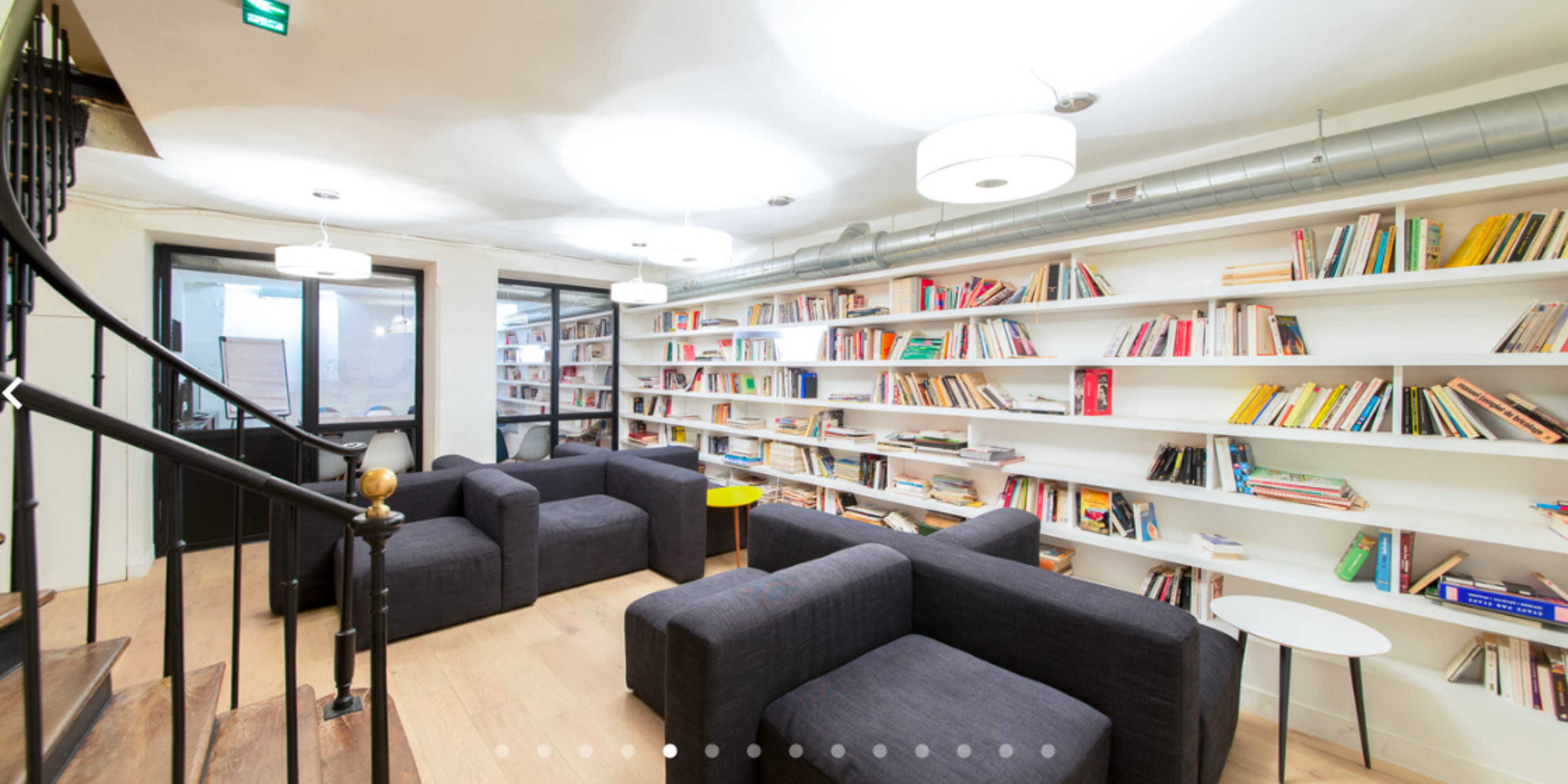 The image shows a modern lounge area with gray seating, a white bookshelf filled with books, and a staircase leading to another level.