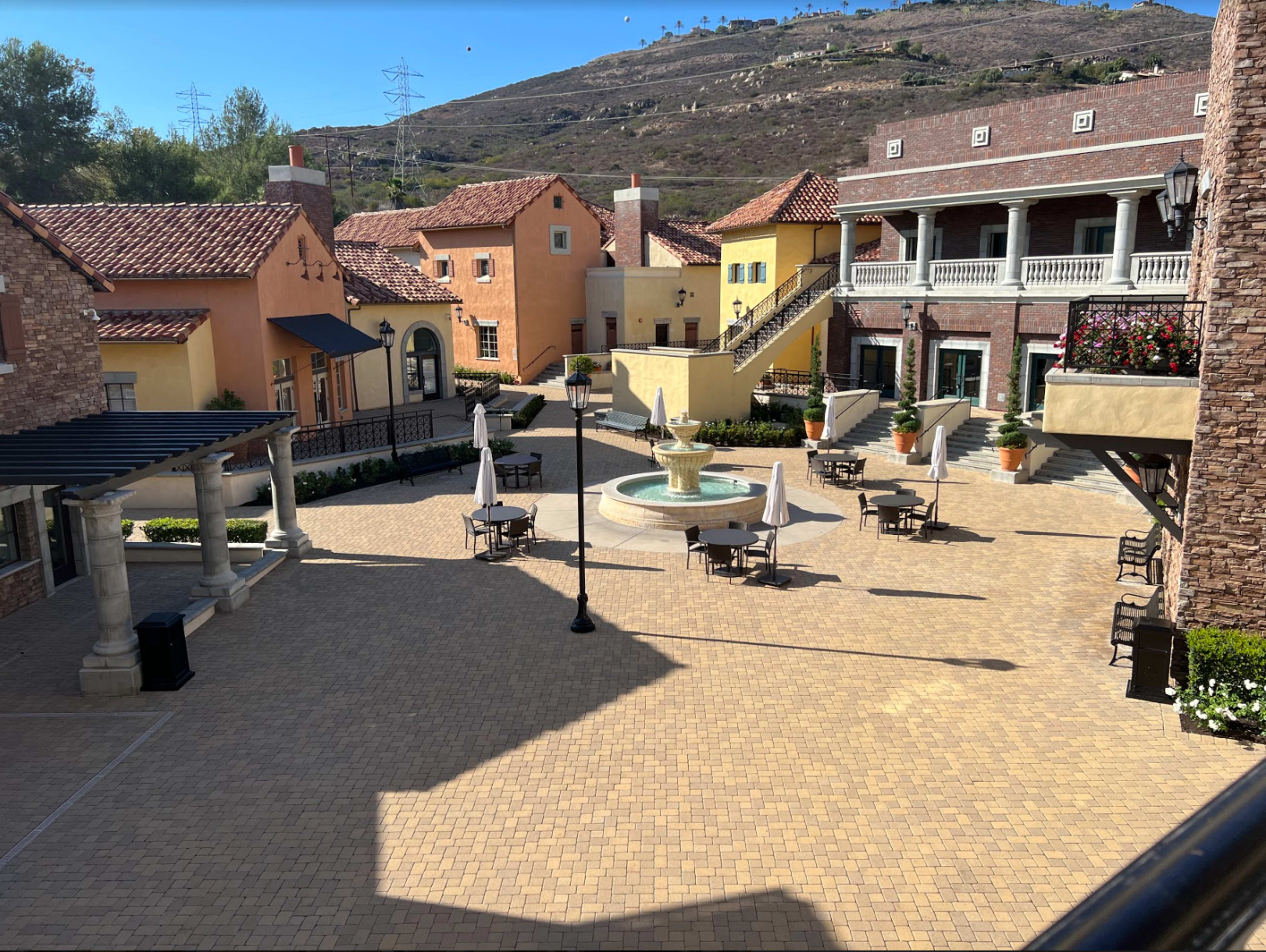 A courtyard with a fountain at the center, surrounded by several buildings featuring a mix of architectural styles and outdoor seating areas.