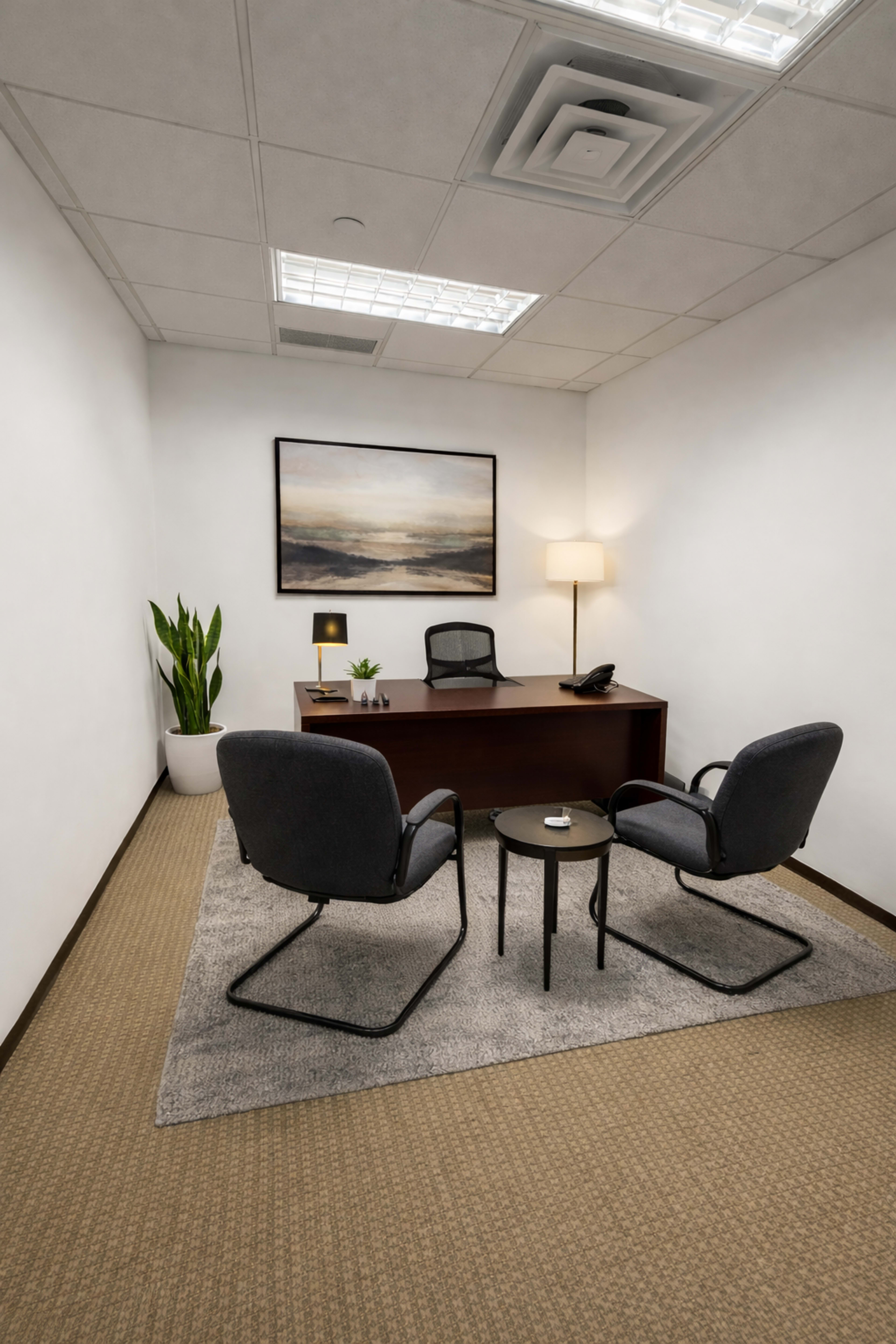 The image shows a small office with a desk, two chairs, a table, and a plant, complemented by a light fixture and a framed landscape painting on the wall.