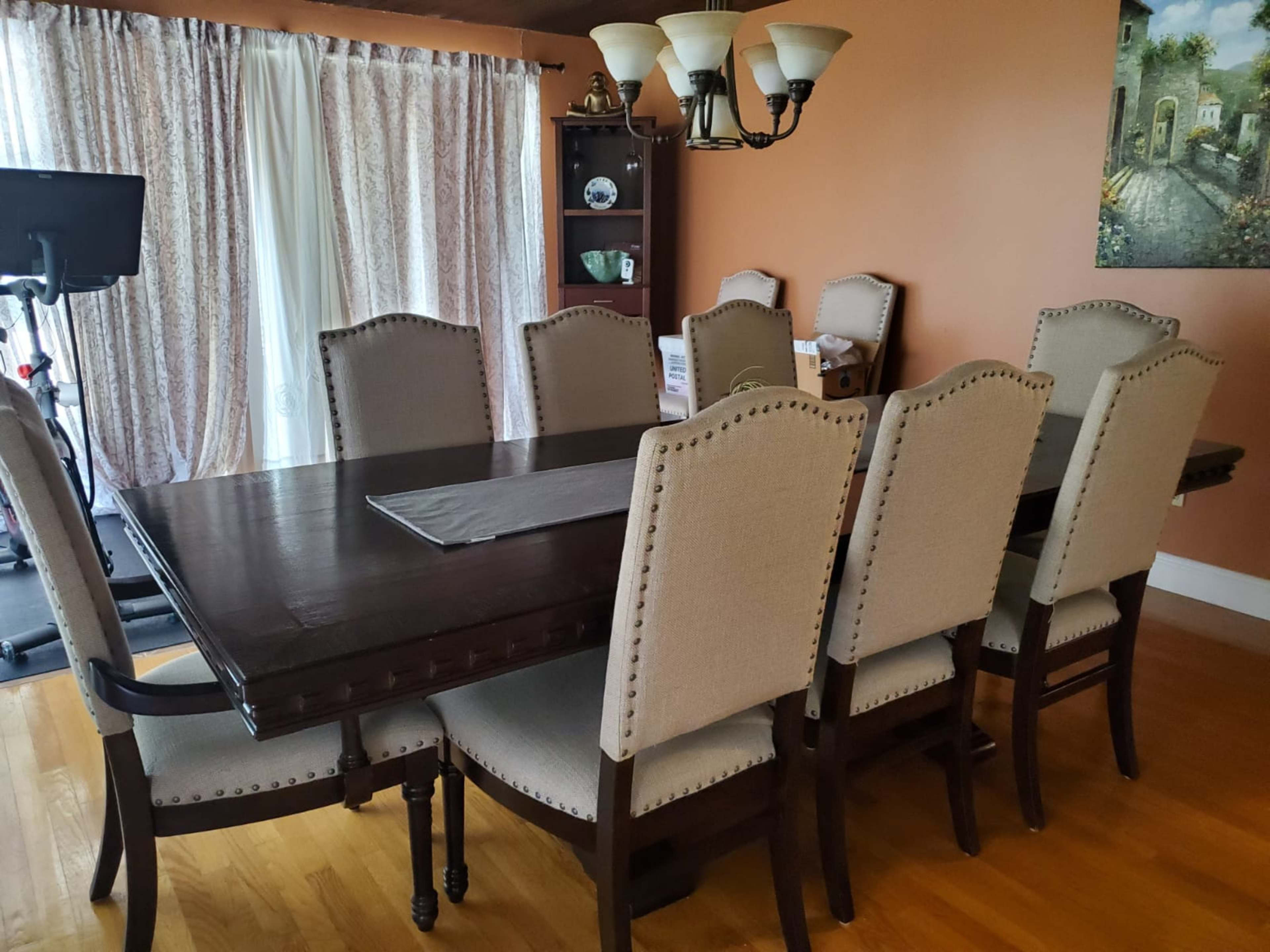 A large wooden dining table with eight upholstered chairs is set against a backdrop of window curtains and a side cabinet in a warmly colored dining room.