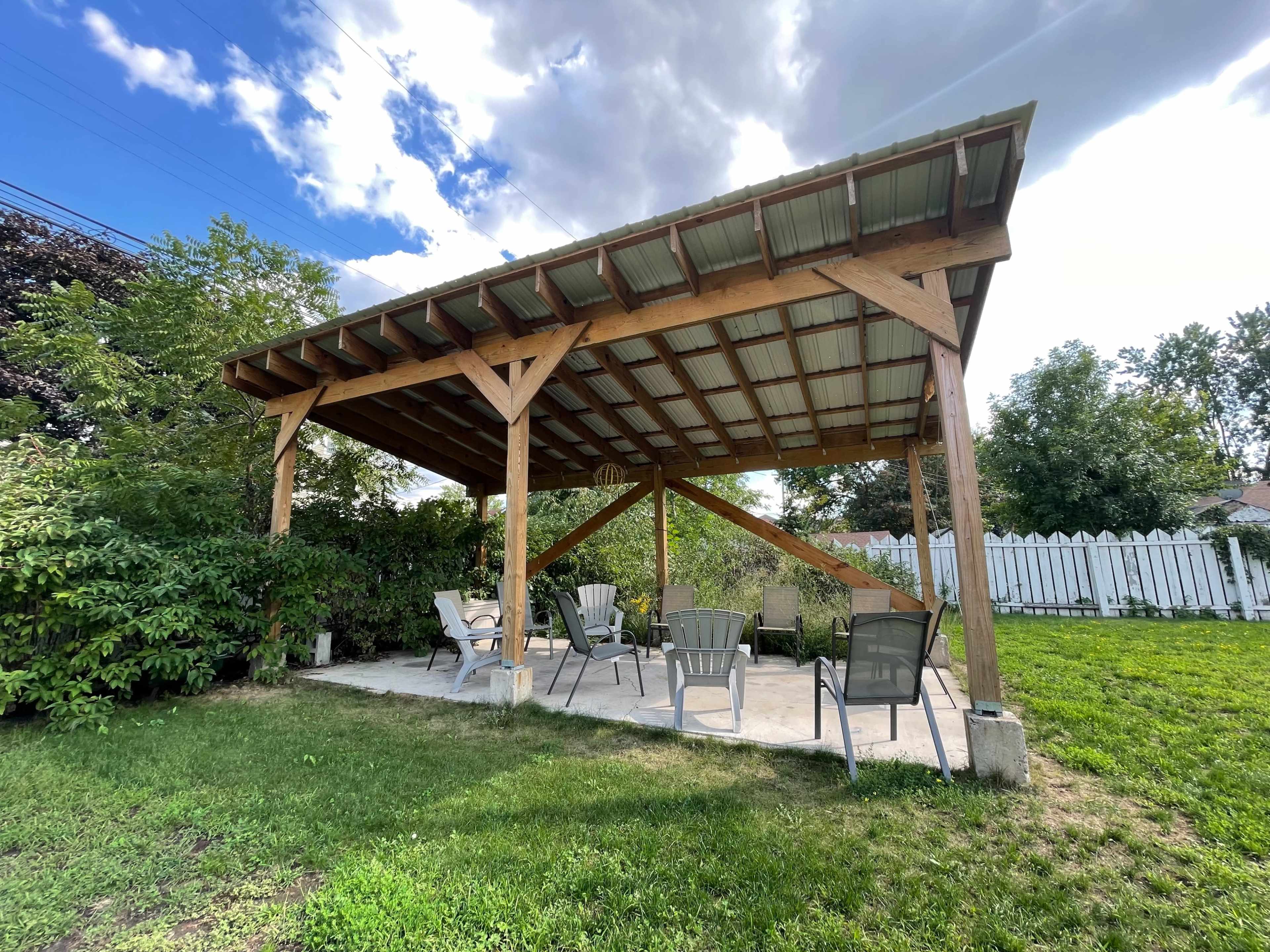 A wooden pavilion with a metal roof stands on a concrete patio, surrounded by greenery and facing a white picket fence.