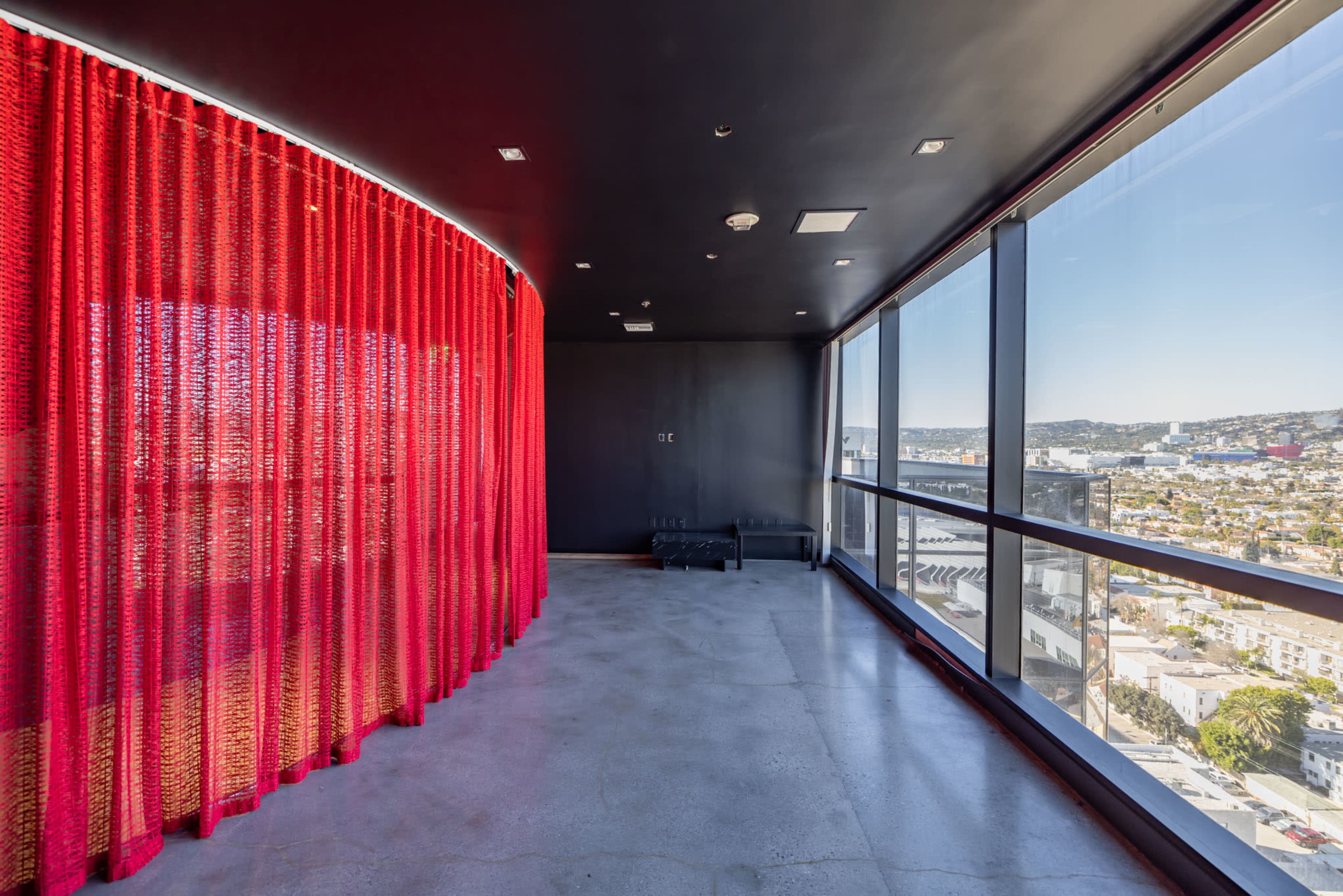 The image shows a modern room with a red curtain, large windows revealing a city view, and concrete flooring.