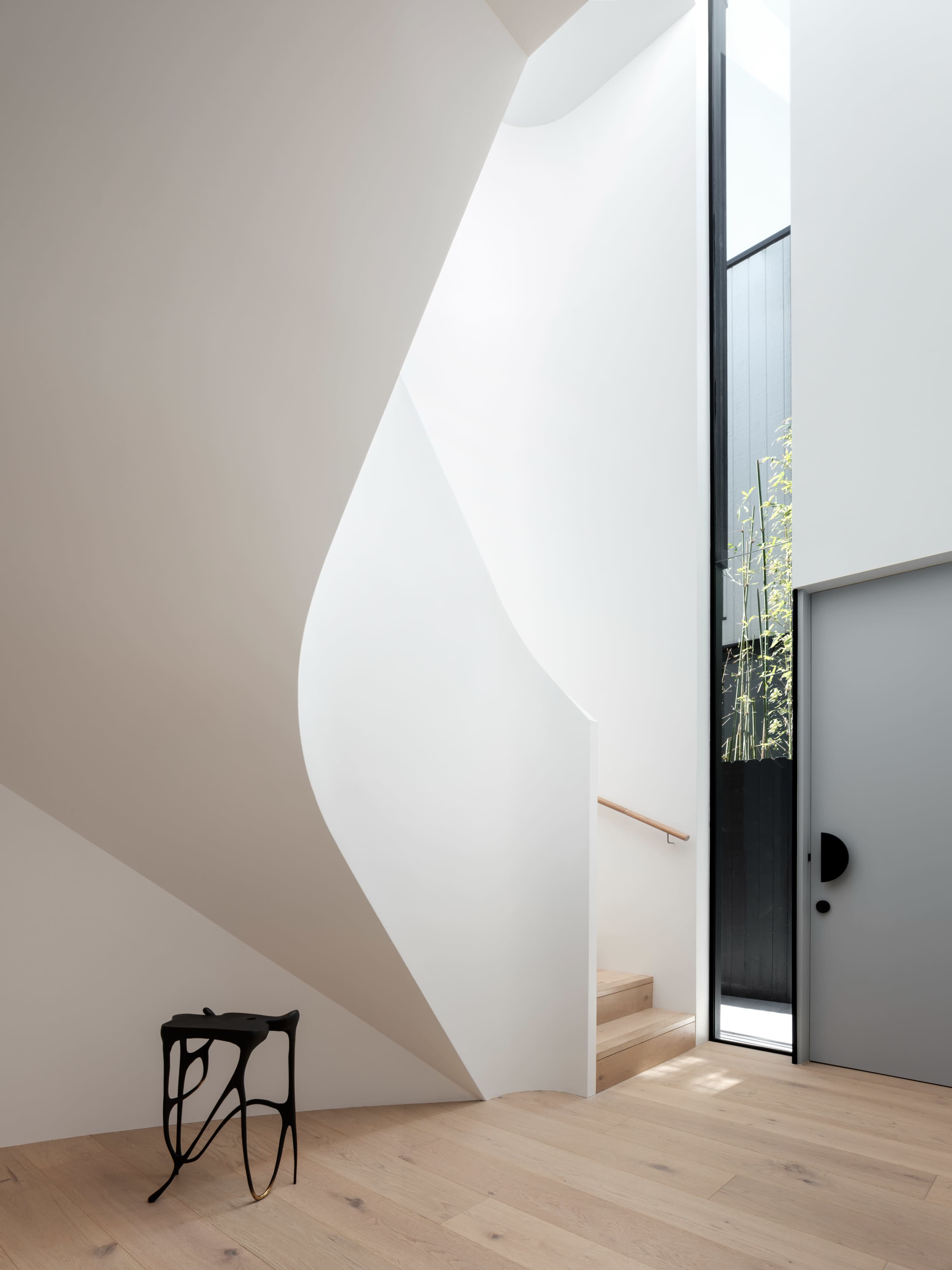 A sleek, modern interior features a curved staircase ascending beside a large glass window that allows natural light to enter.