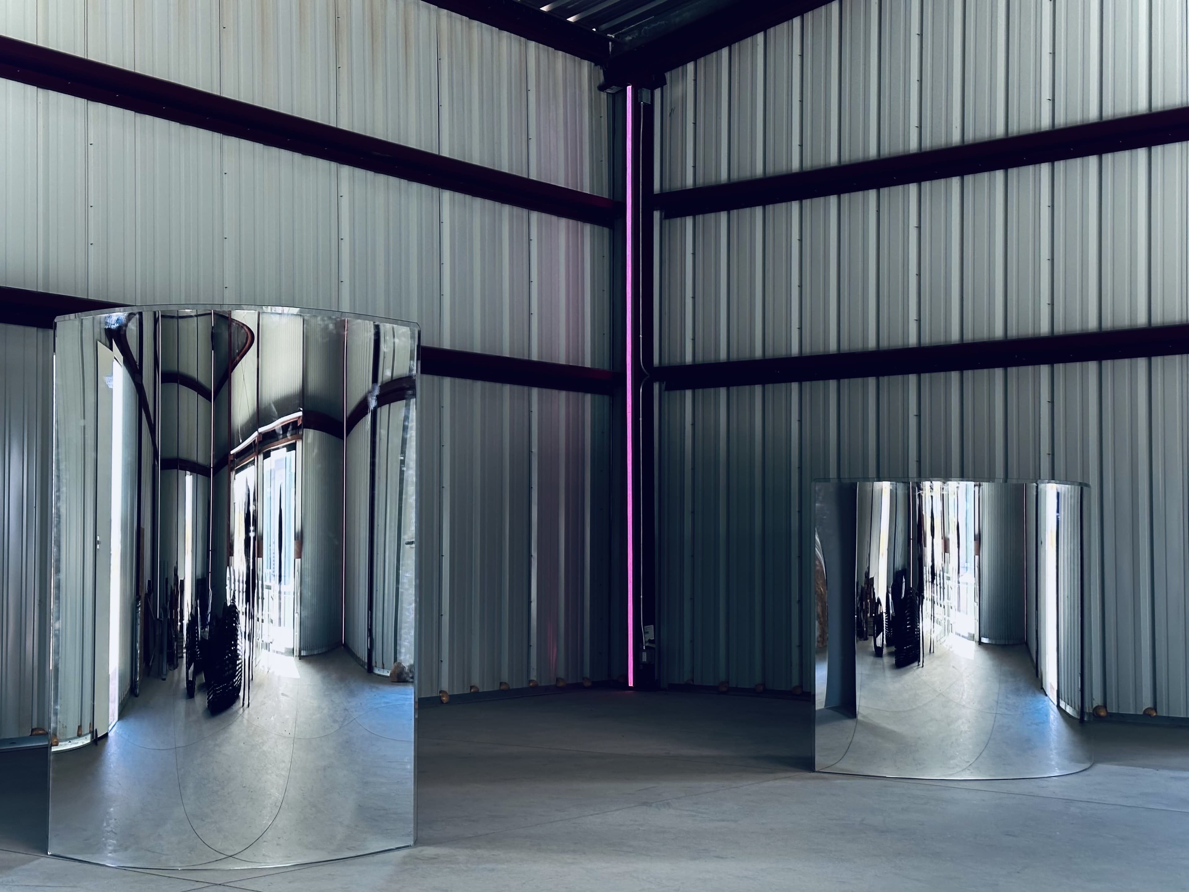 The image shows two large, reflective cylindrical sculptures positioned in a spacious industrial warehouse with corrugated metal walls.
