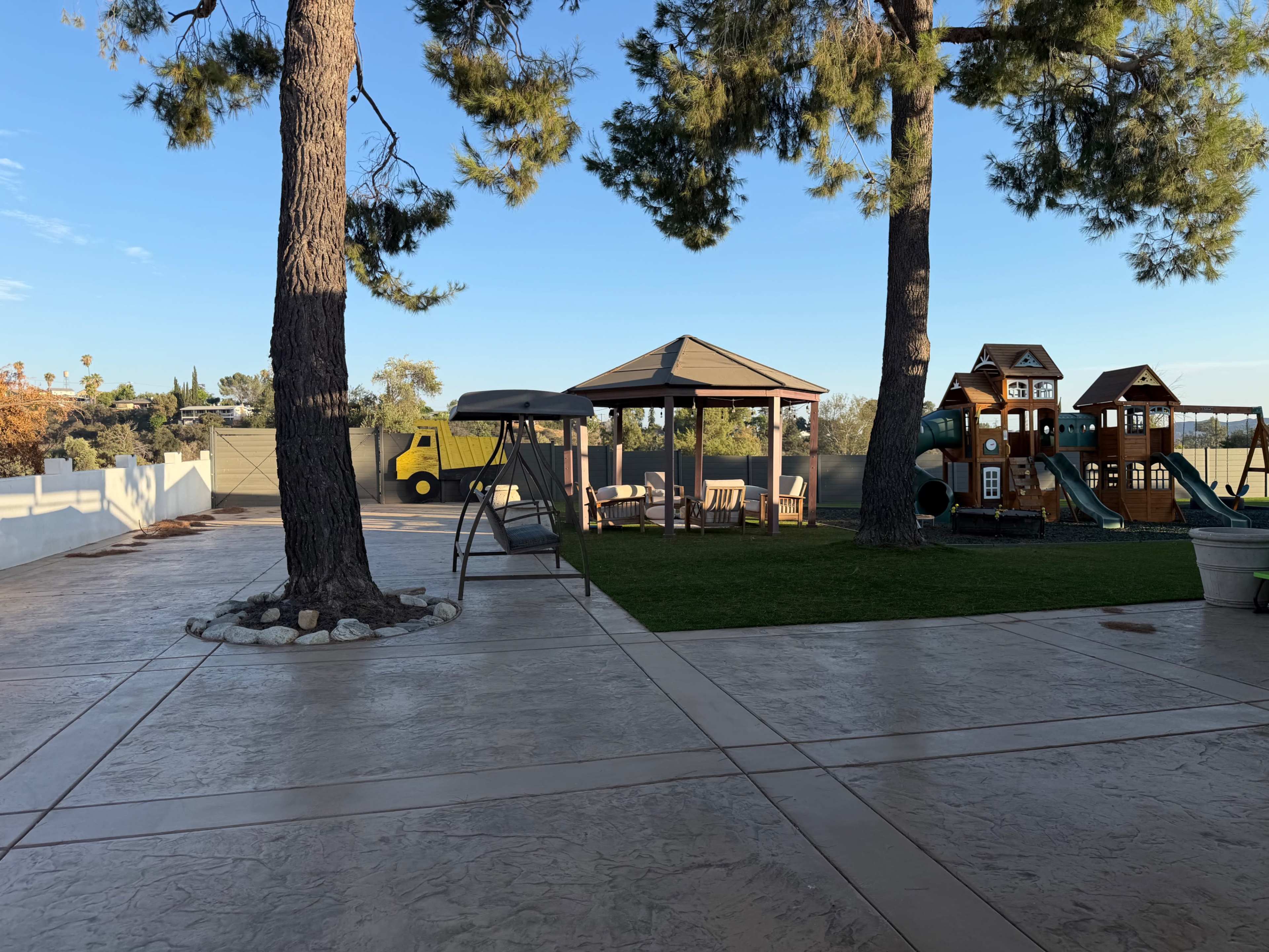 The image shows a spacious outdoor area with a gazebo, a playground set, and a swing beside two tall trees on a sunny day.