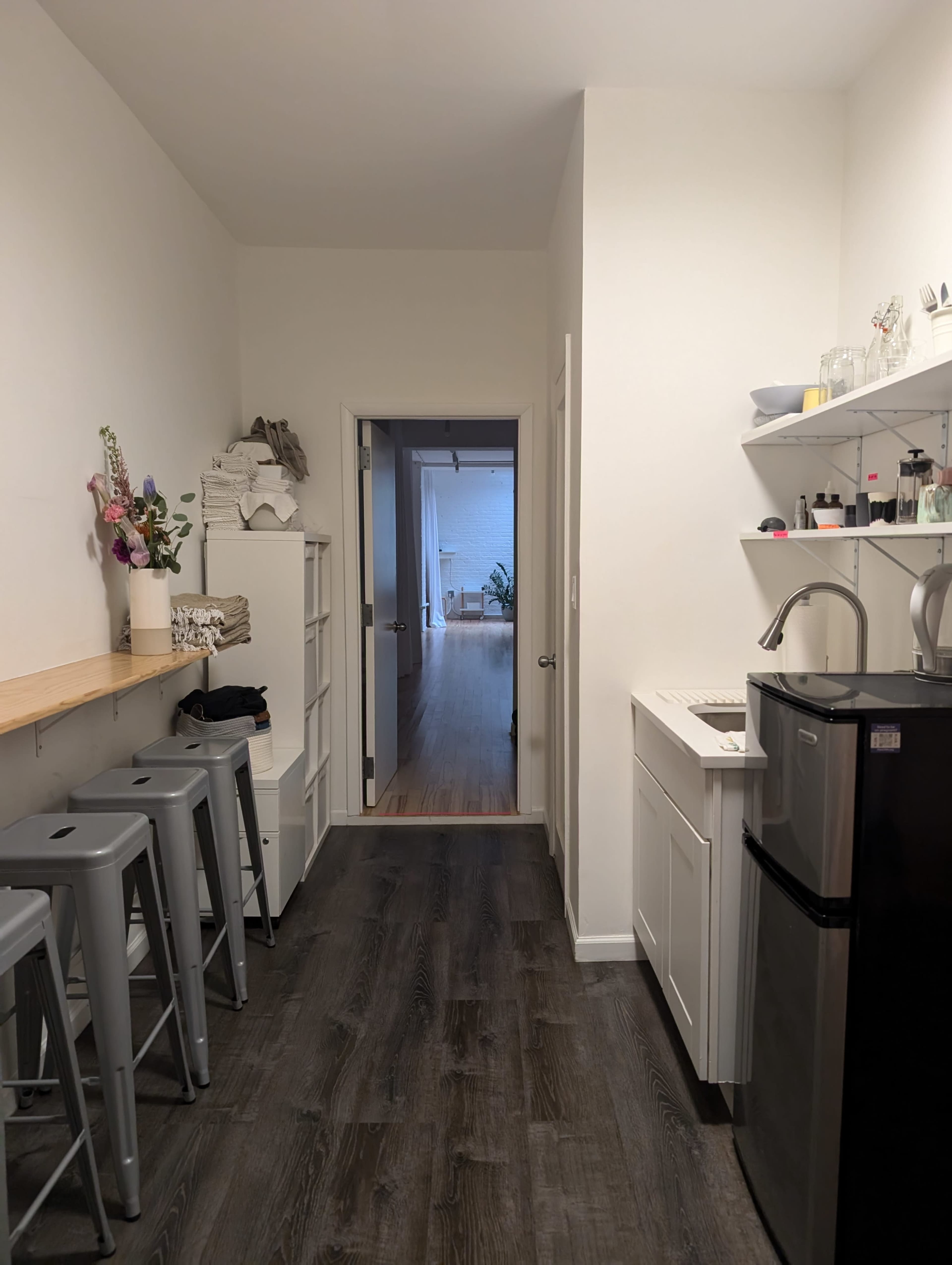 A narrow kitchen hallway features a row of bar stools, shelves with various items, a small refrigerator, and an open doorway leading to a light-filled living space.