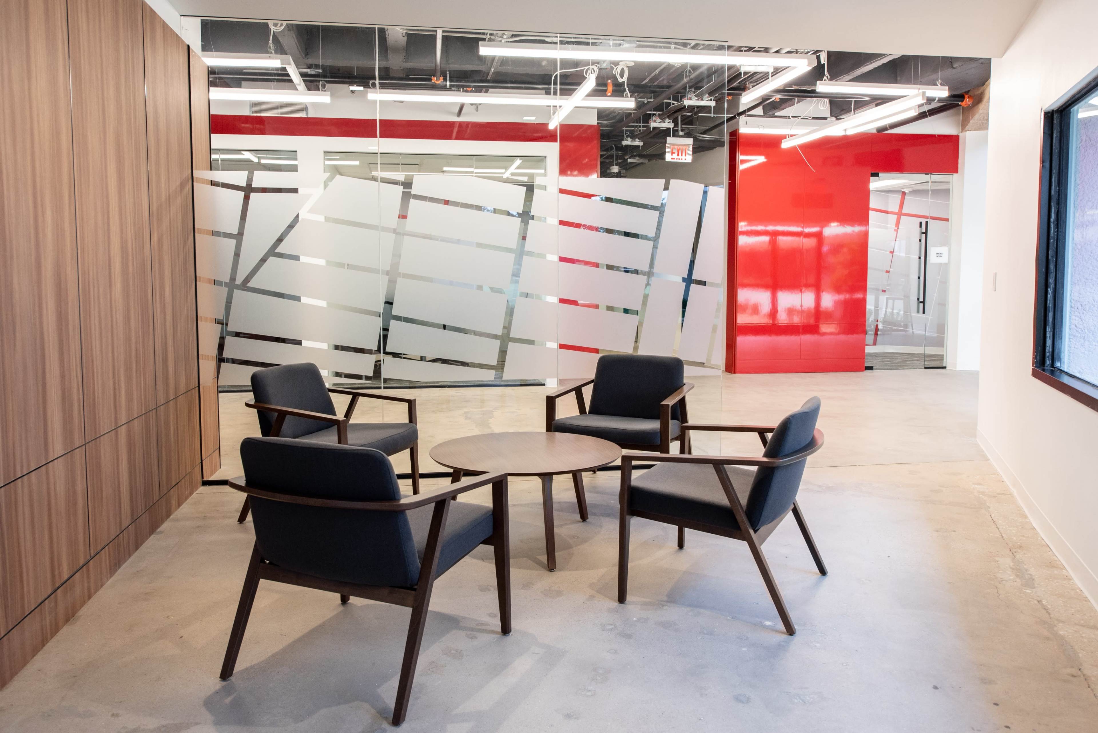 A modern office space with a round table surrounded by four black chairs, adjacent to a wall featuring frosted glass panels and red accents.