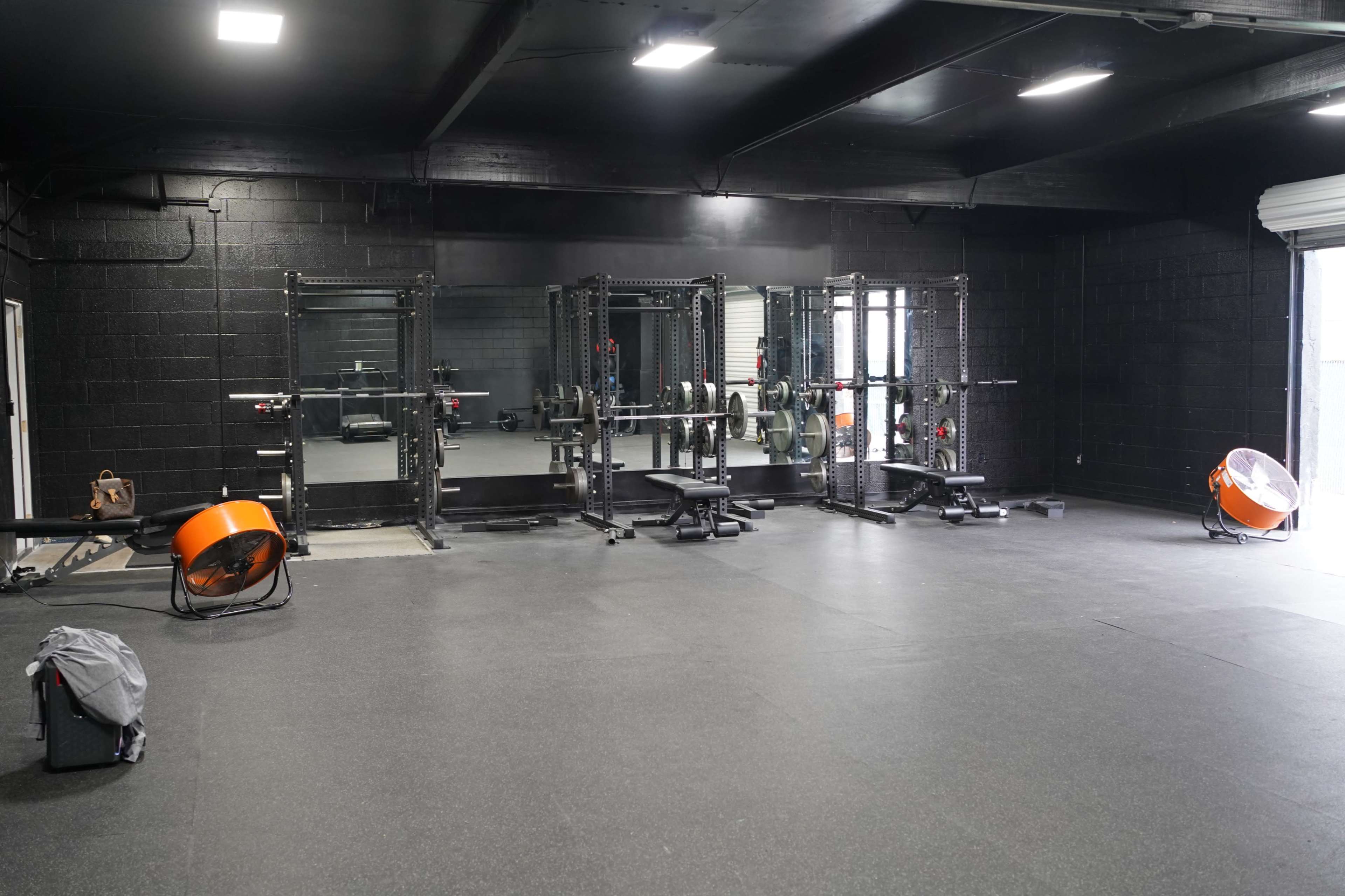 A spacious gym interior with multiple weightlifting rigs, a large fan, and rubber flooring.